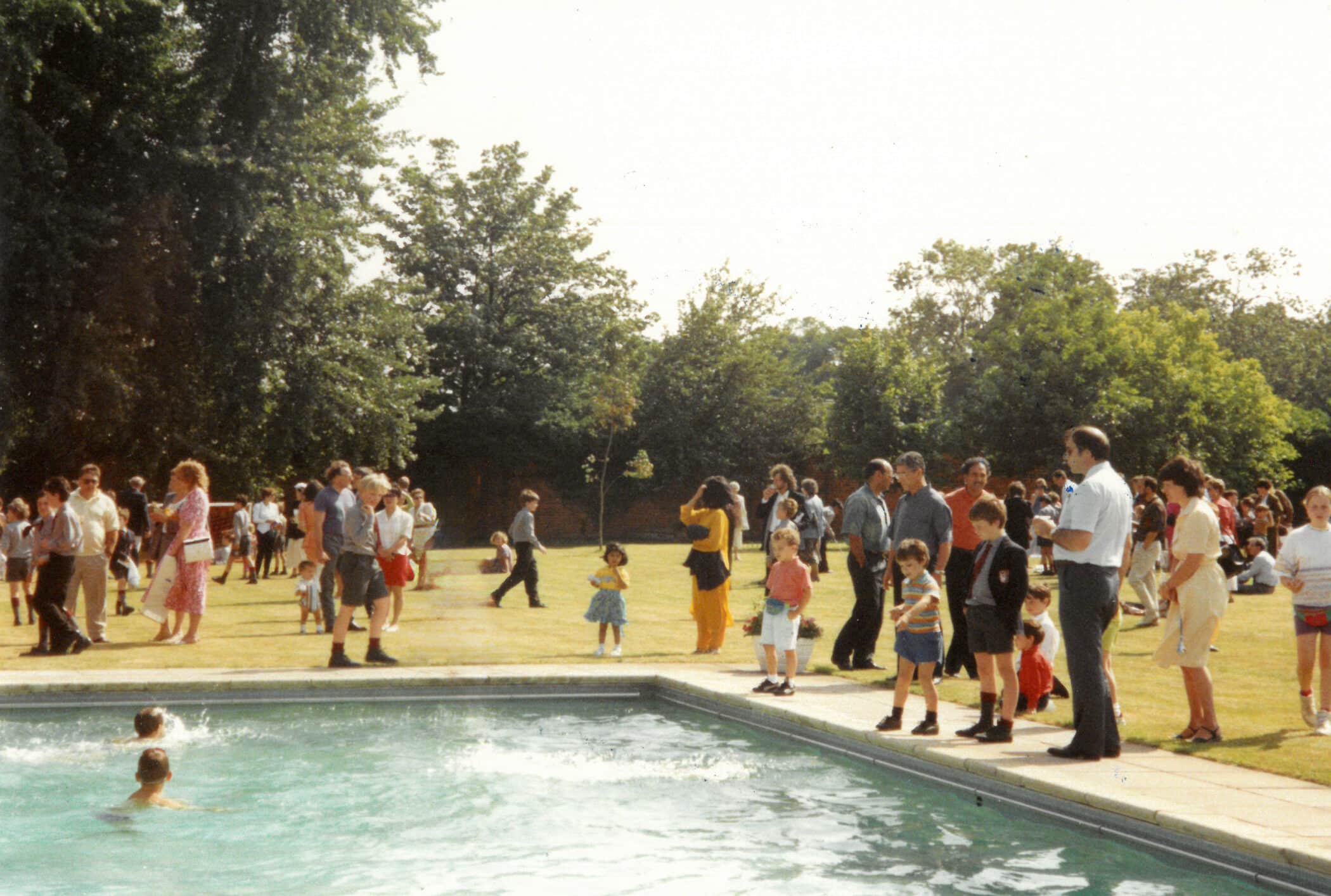 A group of adults and children stand around an outdoor swimming pool, watching two children swim. More people are scattered on the grassy area nearby, surrounded by trees on a bright, sunny day.