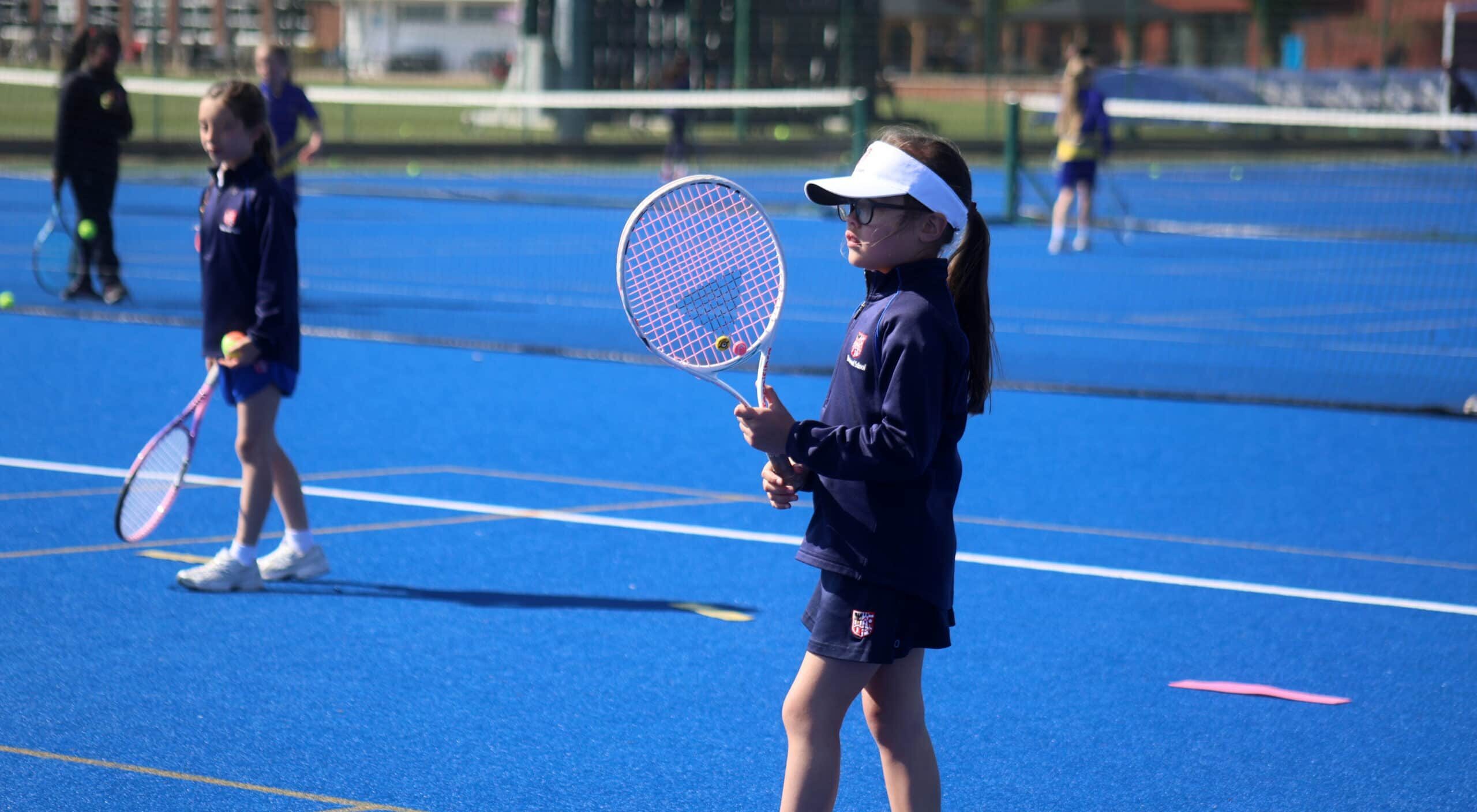 A young girl in sports kit and a visor holds a tennis racquet on a blue outdoor court, preparing to play. Other children with racquets stand nearby, reflecting the vibrant spirit of prep school education.
