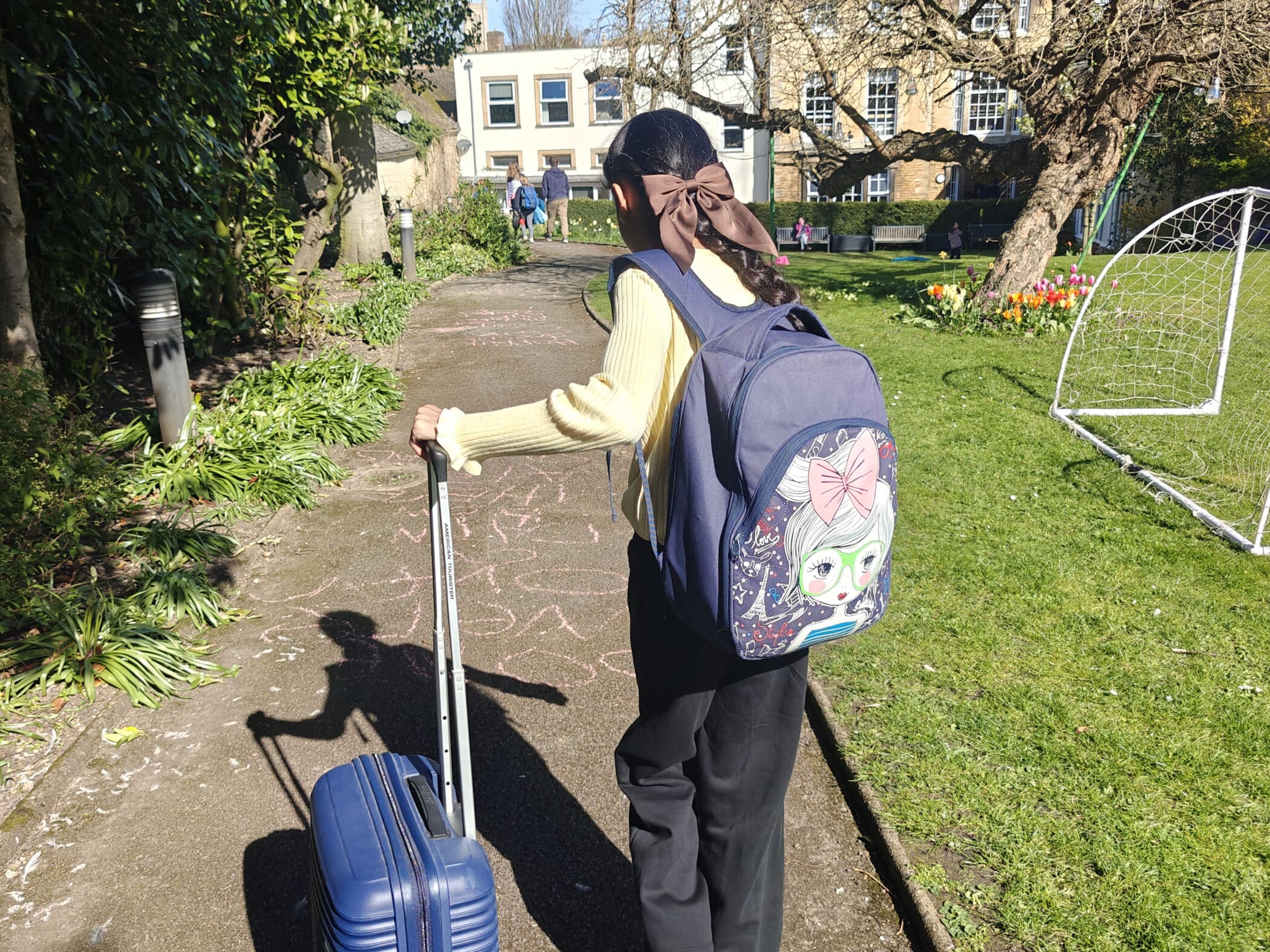 A child with a large rucksack and a rolling suitcase walks along a sunlit path in a garden, with chalk drawings on the ground and buildings, grass, flowers, and a football goal nearby.