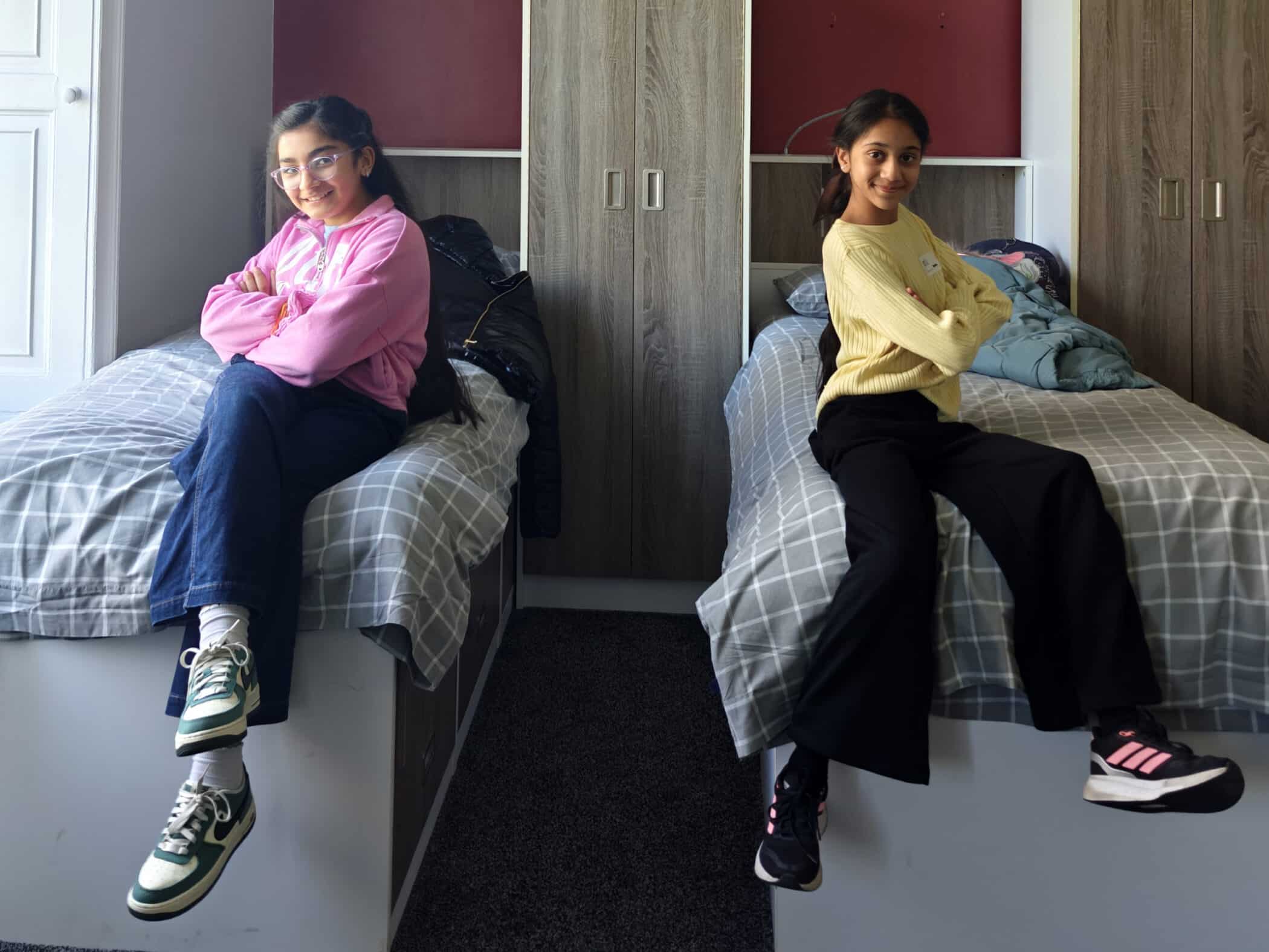 Two girls sit on separate beds in a bedroom, each smiling with arms crossed. One wears a pink top and trainers, the other a yellow jumper and black trousers. Jackets are laid on the beds, and wardrobes are behind them.