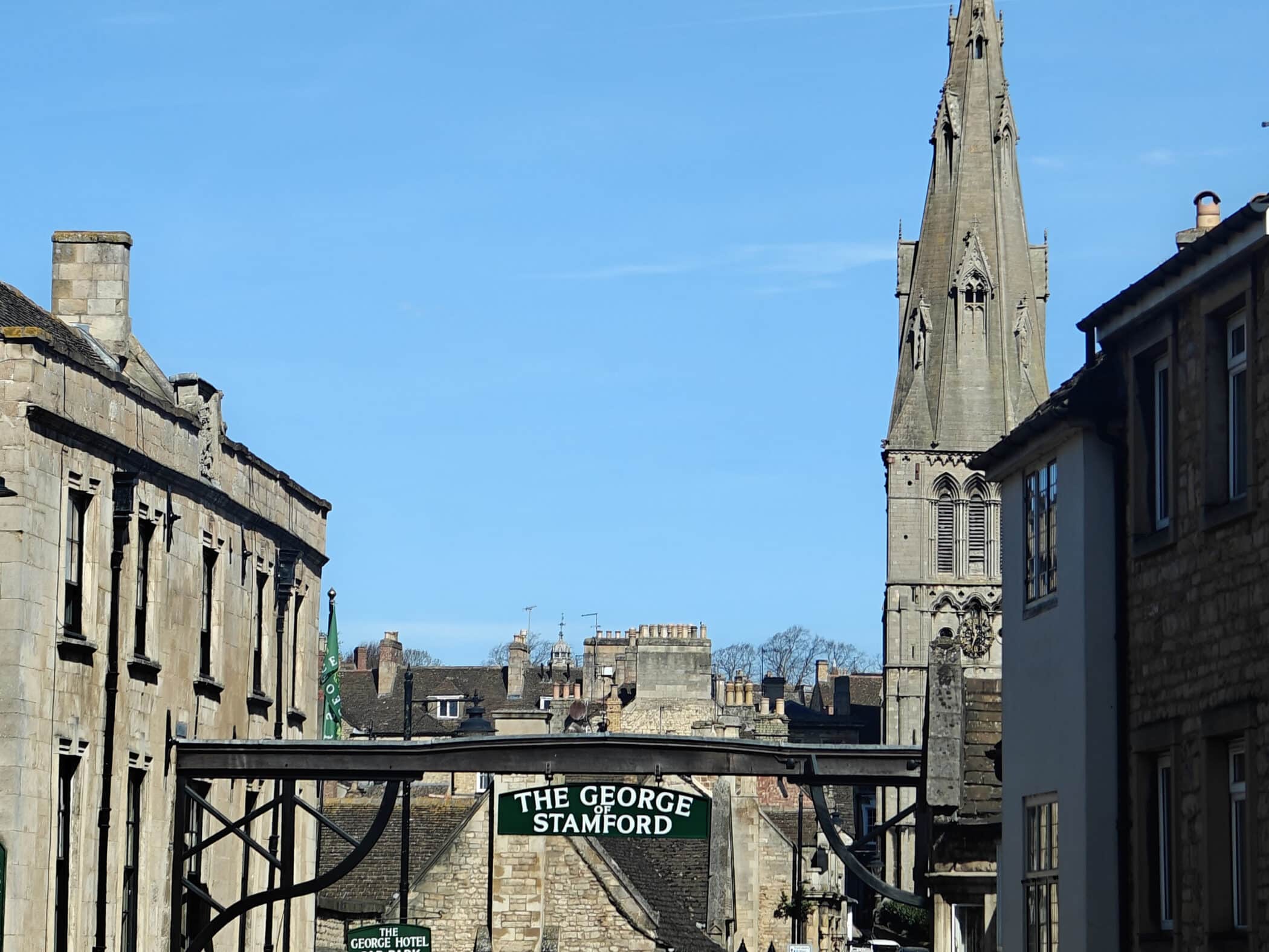 View of a historic town street with stone buildings, a sign reading The George Stamford, and a tall church spire rising in the background against a clear blue sky.
