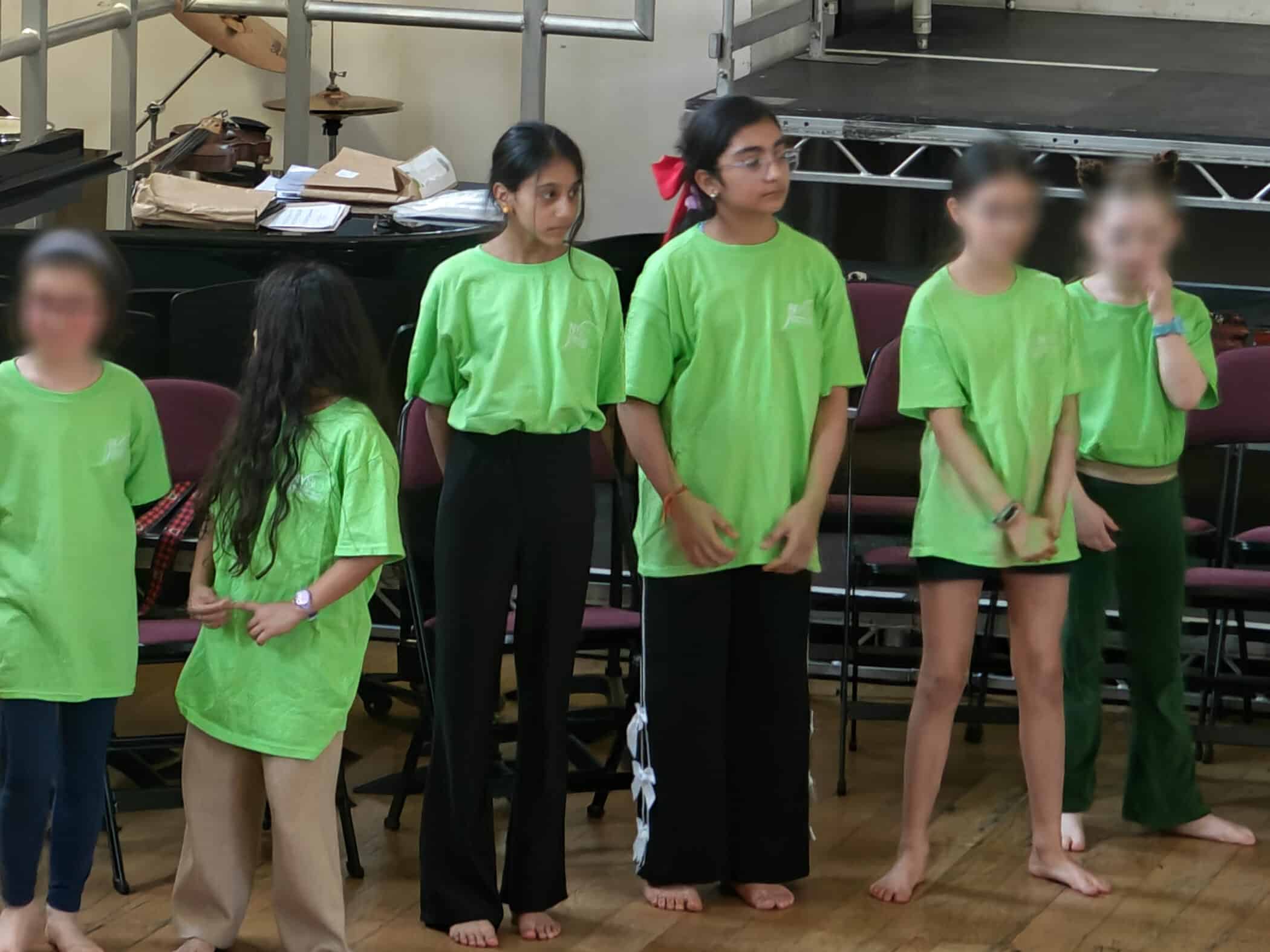 A group of five barefoot children stand indoors, all wearing bright green T-shirts. Some children have long hair, and they are in front of a row of chairs and stage equipment. Two faces are clearly visible; three are blurred.