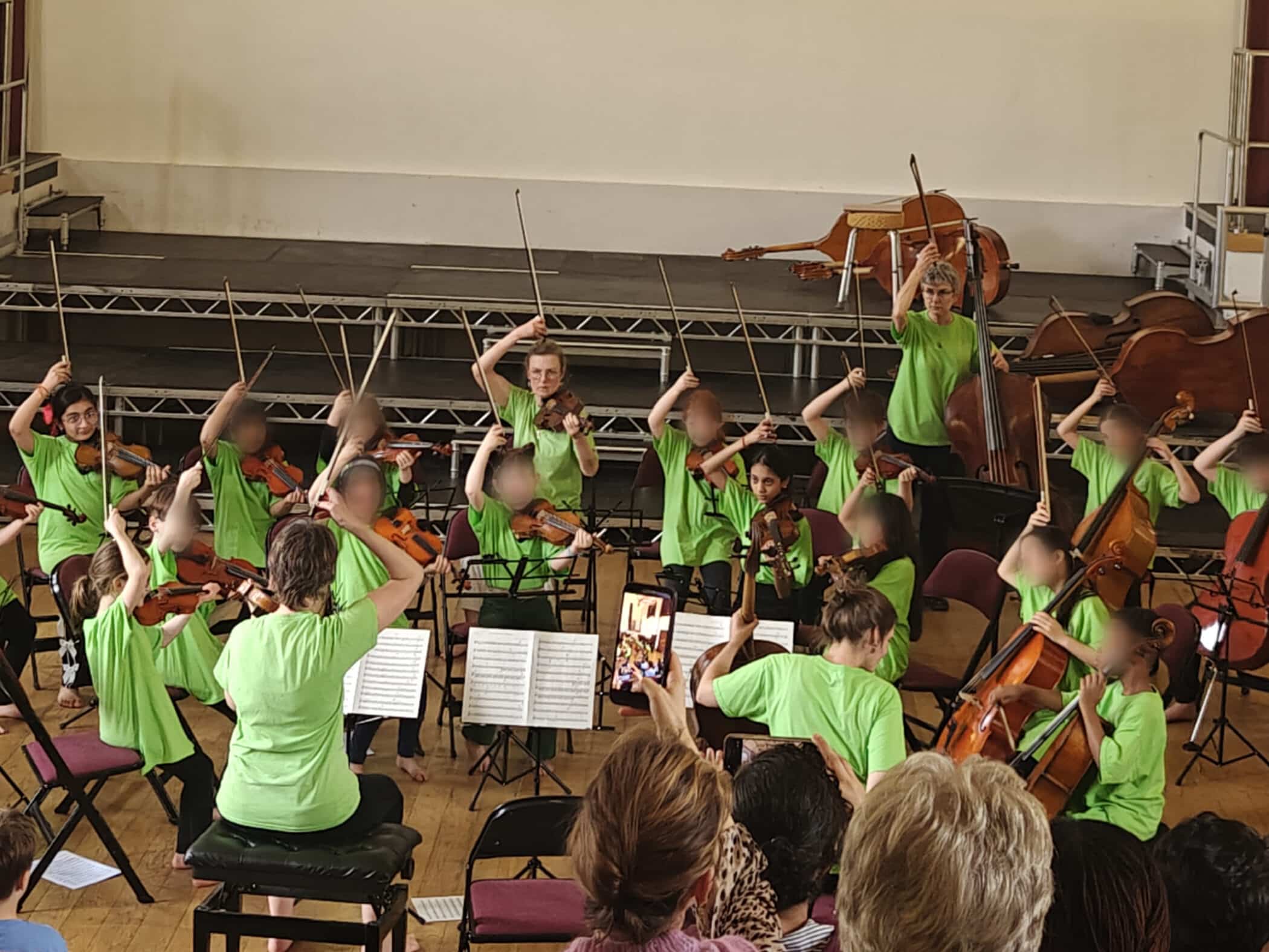A children's orchestra in bright green shirts performs on stage, raising their bows in the air. A conductor leads from the front, while an audience watches and someone records the performance on a mobile.