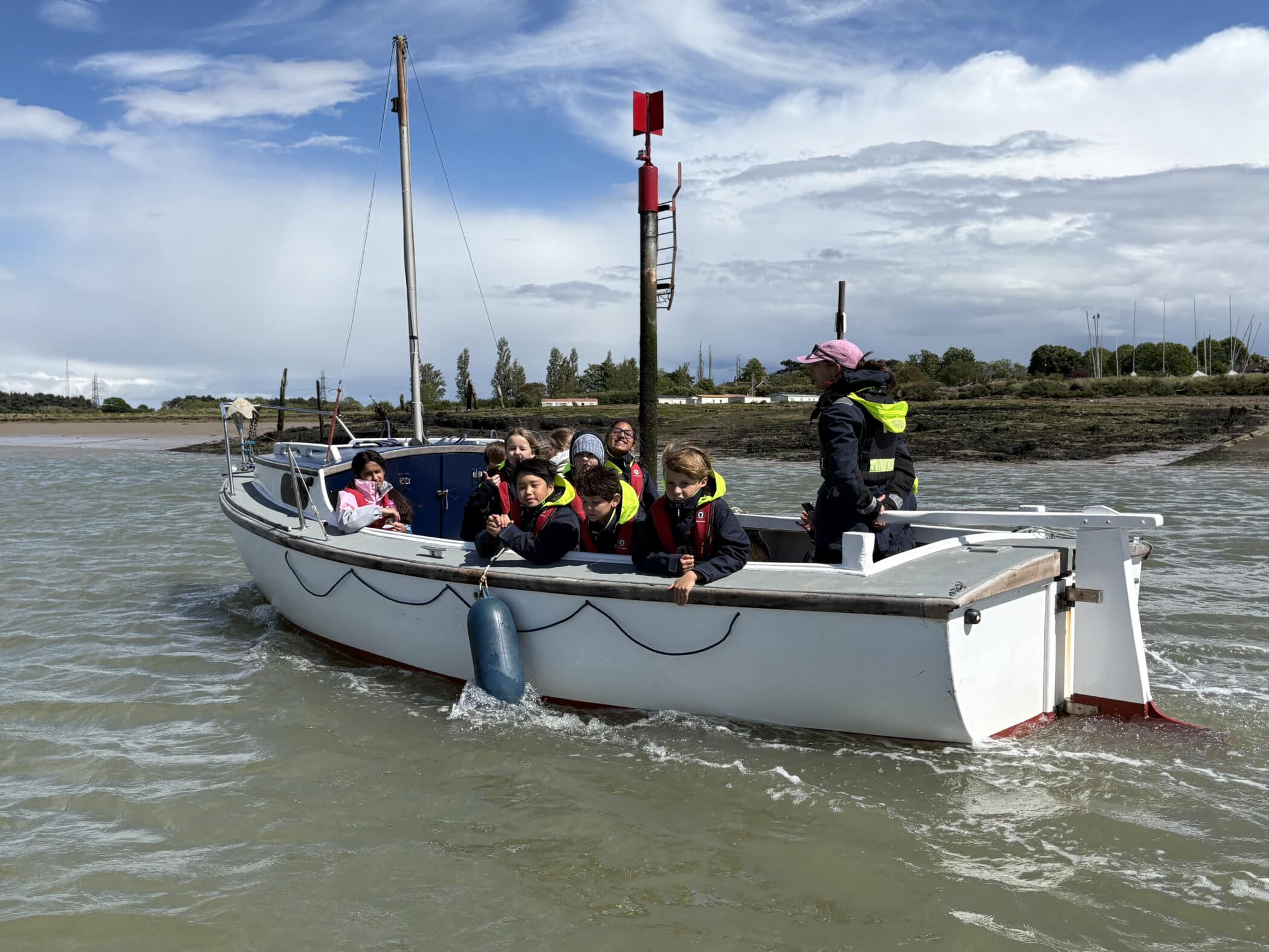 A group of children and an adult wearing life jackets ride a white boat on a cloudy day, with land, trees, and boats visible in the background. The water is choppy and everyone appears to be enjoying the outing.