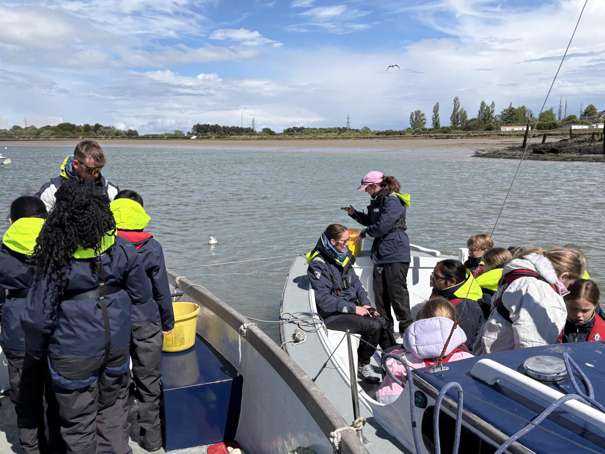 A group of children and adults wearing life jackets sit and stand on a boat moored on a river under a partly cloudy sky, with trees and buildings visible on the riverbank in the background.