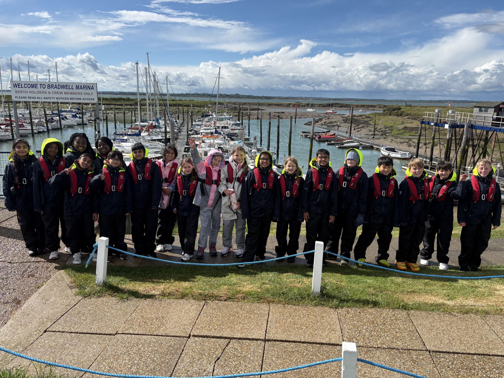 A large group of children in matching jackets and life vests stand in a queue by a marina with boats and sailing boats in the background, under a partly cloudy sky.