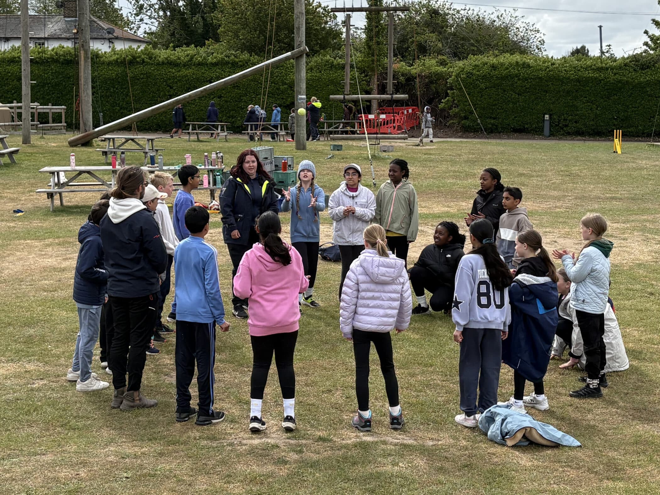 A group of children and two adults stand in a circle on a grassy field, some clapping and talking. Picnic tables, playground equipment, and trees are visible in the background on a cloudy day.