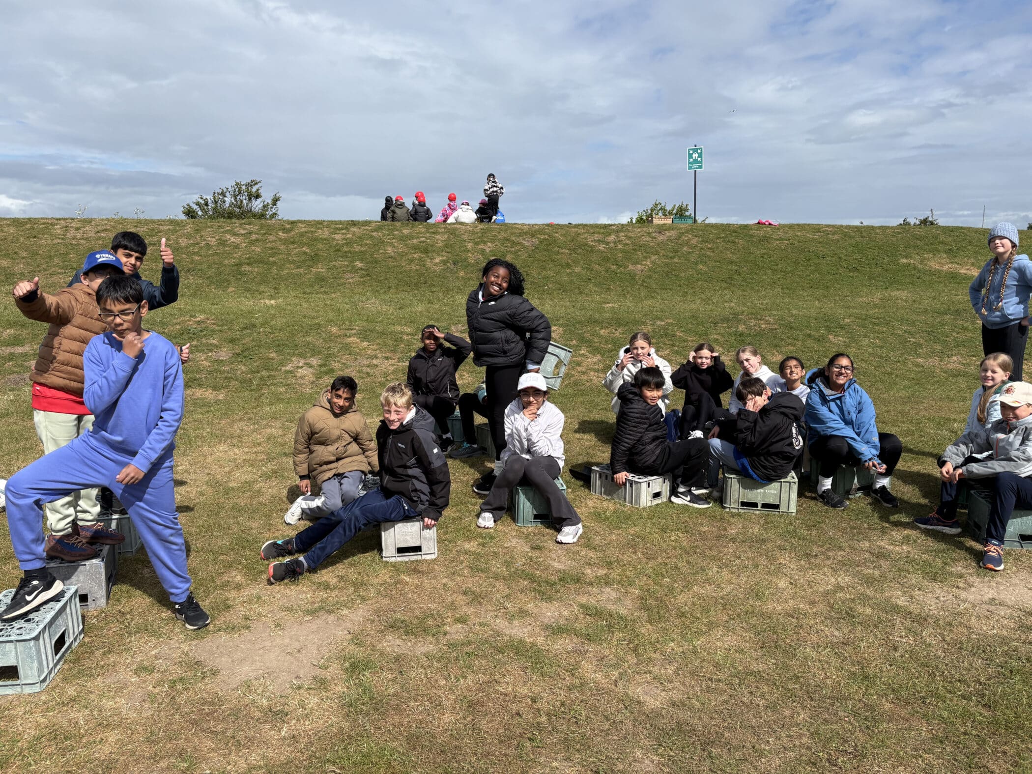 A group of children sit and pose on plastic crates outdoors on a grassy field, smiling and making playful gestures. A few people sit on a hill in the background under a partly cloudy sky.