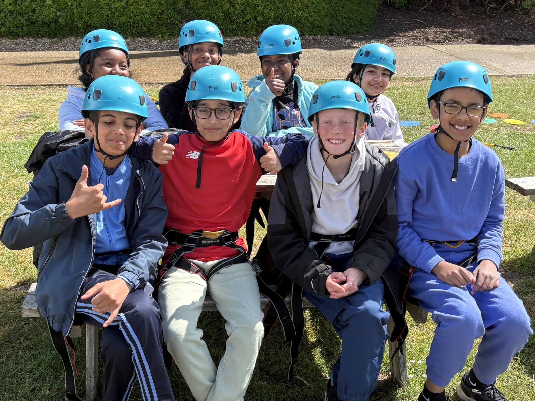 A group of smiling children wearing blue helmets and outdoor clothing sit together on a bench outside, posing for a photo on a sunny day. Some are giving thumbs up or peace signs, looking excited and happy.