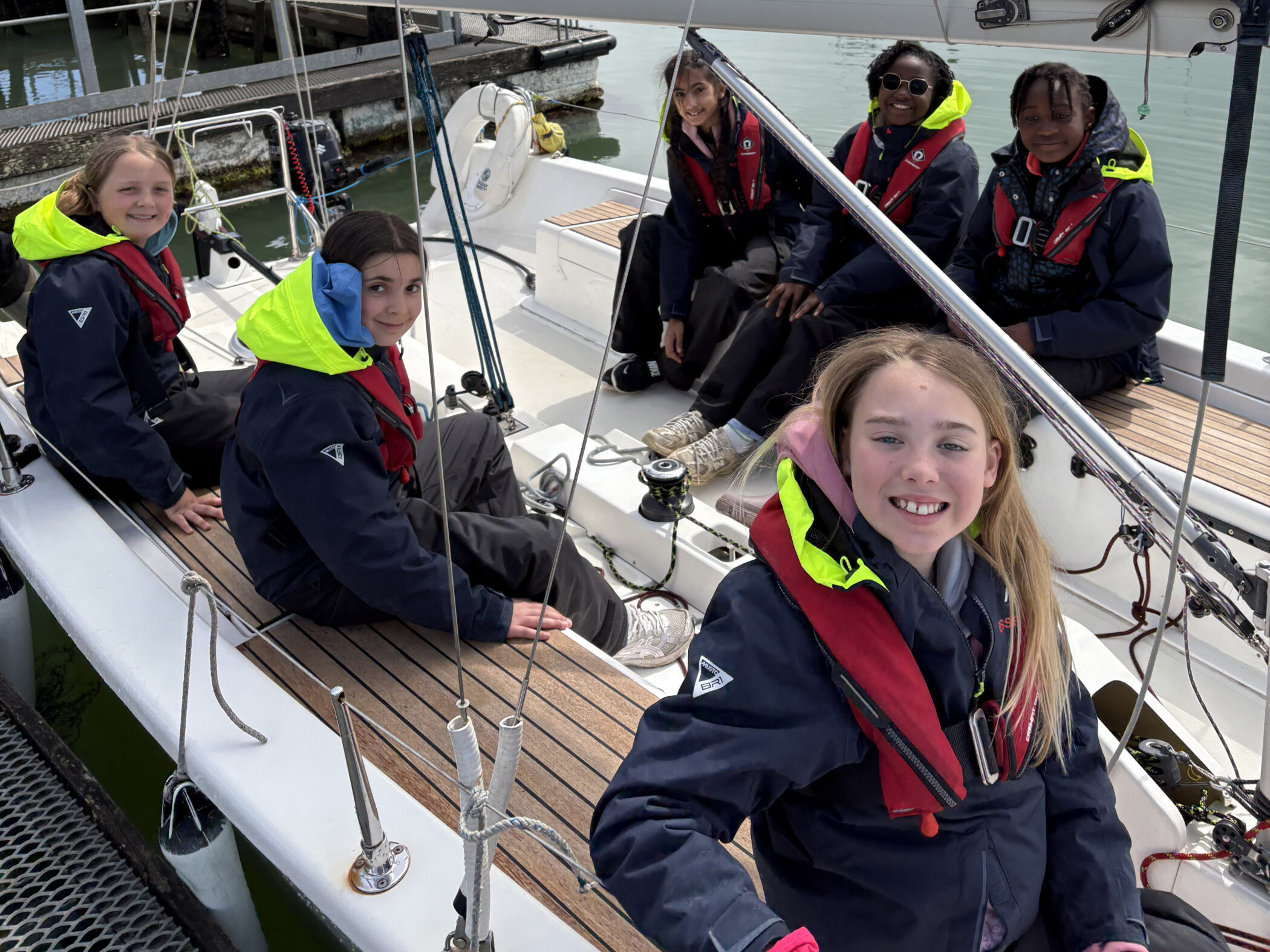 Five smiling girls in sailing jackets and life vests sit on a moored sailboat, ready to sail. The water and quay are visible in the background. The girls appear happy and excited, enjoying an outdoor adventure together.
