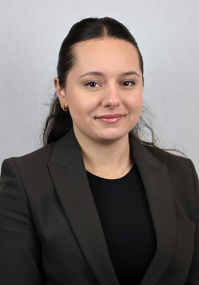 A woman with long dark hair, wearing a dark blazer and black top, is posed against a light grey background. She is facing the camera and smiling softly.
