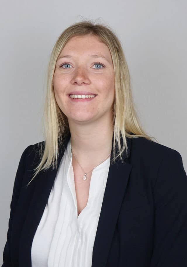 A woman with straight blonde hair is smiling at the camera. She is wearing a dark blazer over a white blouse and a silver necklace, posed against a plain light grey background.