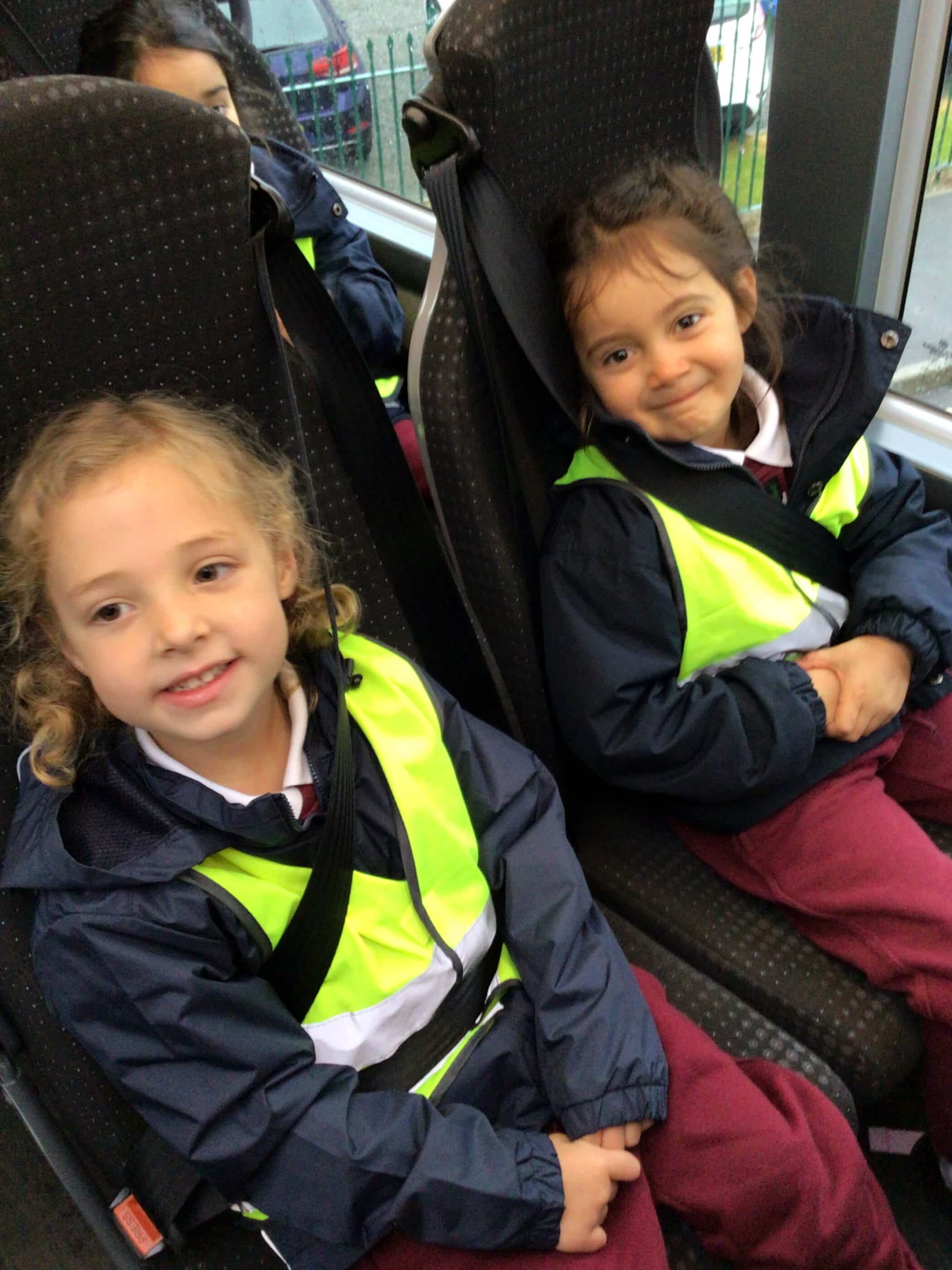 Two young children wearing high-visibility vests and maroon trousers are sitting next to each other on a bus, smiling. They both have seatbelts on and look ready for a journey.