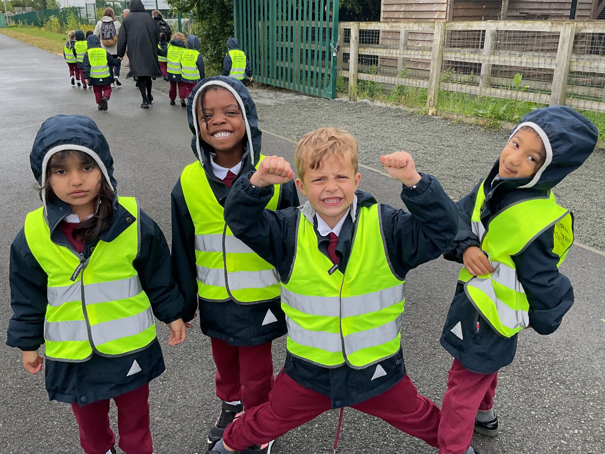 Four young children in matching school uniforms and reflective yellow vests stand outside on a wet day. Two are smiling, one is flexing his arms playfully, and another looks serious. More children and adults walk ahead in the background.