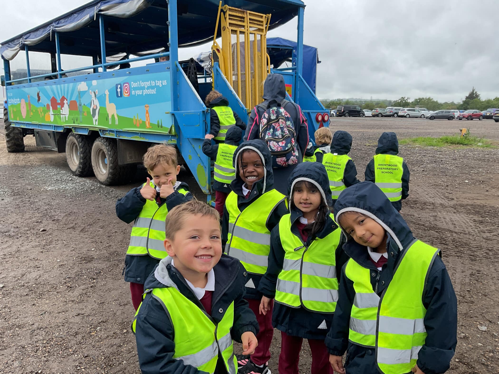 A group of young children wearing yellow high-visibility vests and raincoats stand near a blue lorry trailer. Some smile and give thumbs up while others queue to board the trailer on a cloudy day.