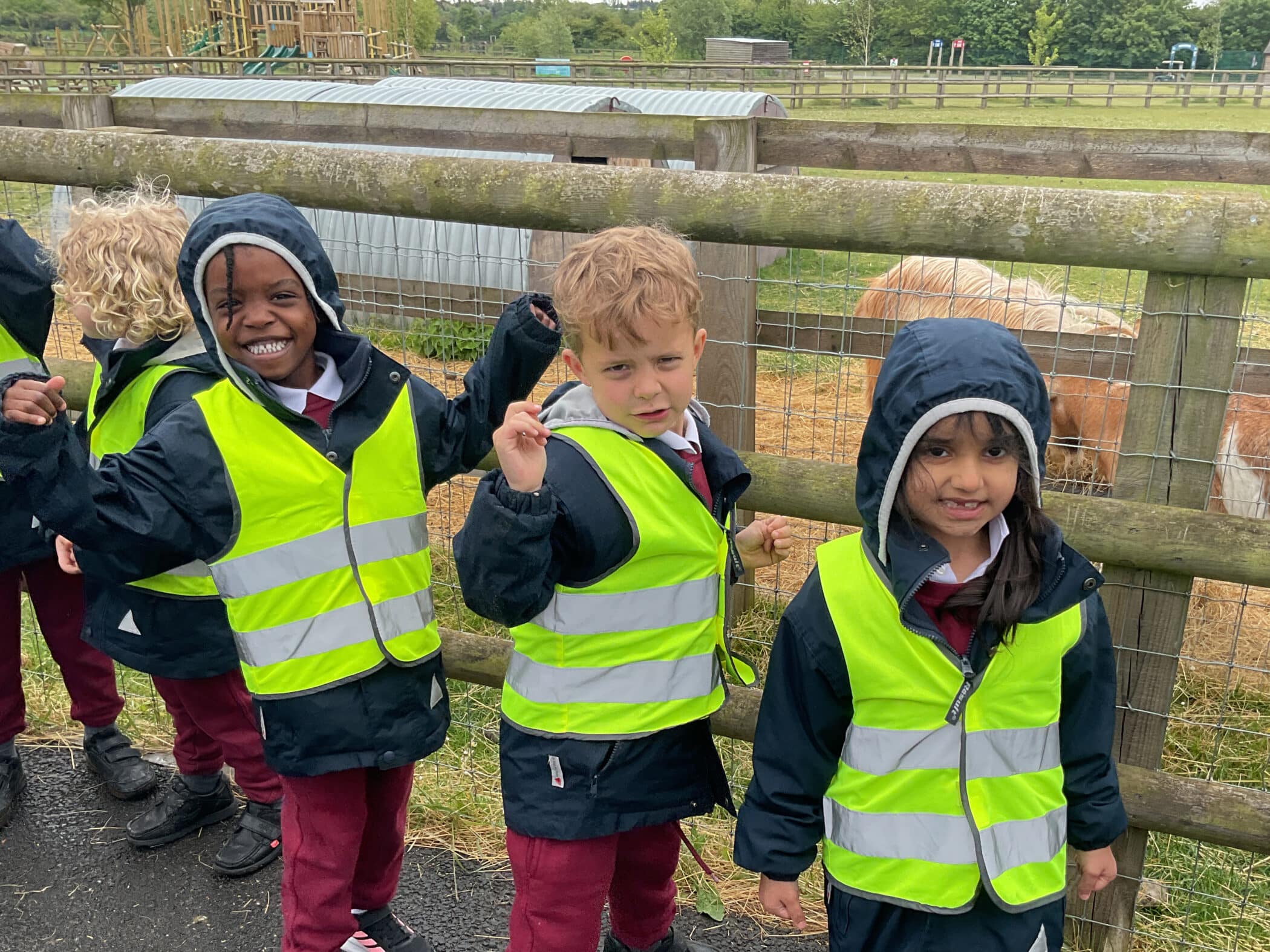 Three young children wearing reflective yellow safety vests and rain jackets stand by a wooden fence at a farm. Two of them smile whilst one has a neutral expression. Ponies and farm structures are visible in the background.