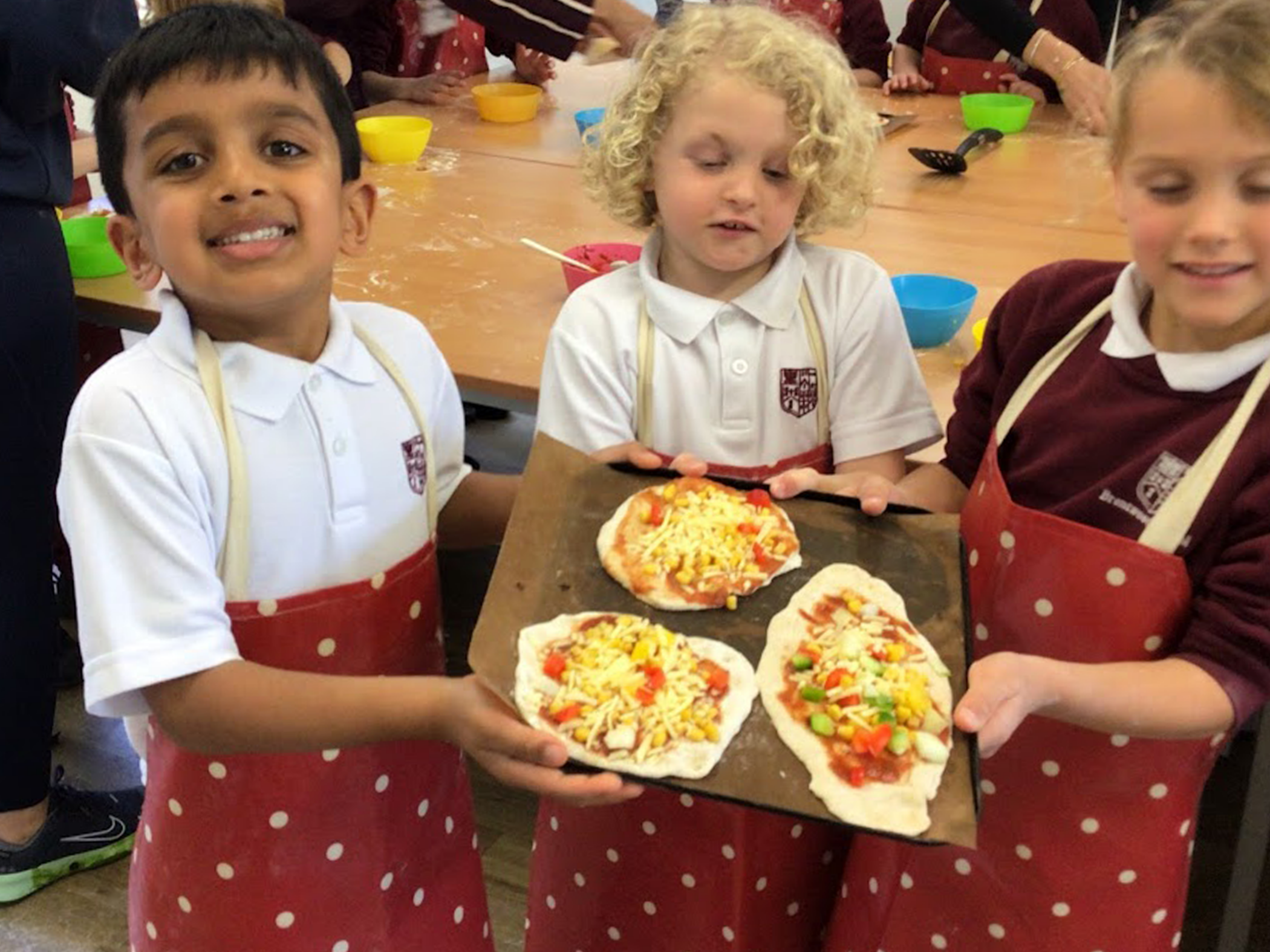 Three young children wearing red polka dot aprons hold a tray with three uncooked homemade pizzas, smiling in a classroom or kitchen setting with other children and colourful bowls in the background.