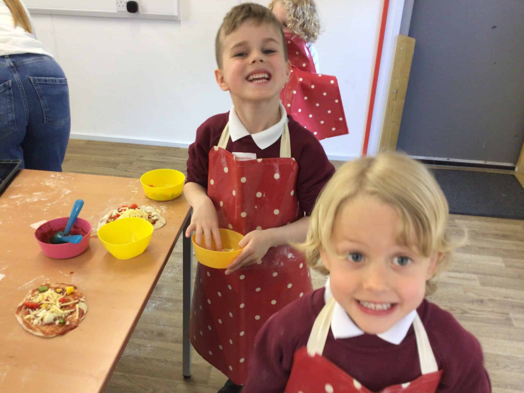 Two young children wearing red polka dot aprons smile at the camera whilst making pizzas. They stand at a table with bowls of ingredients and small pizzas. Another child and an adult are in the background.