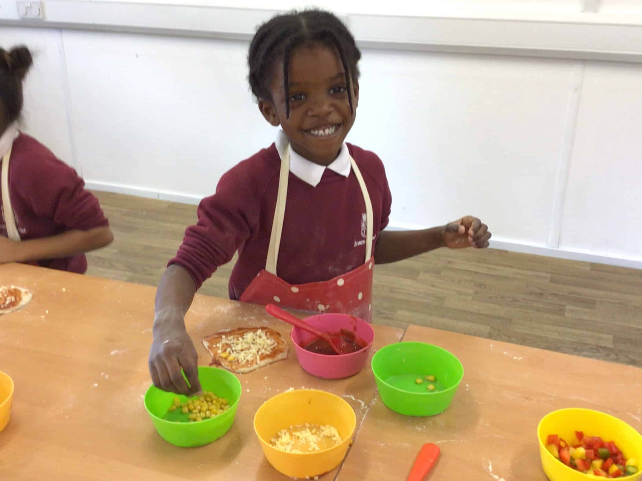 A smiling child in a red pinafore and school uniform prepares food at a table, placing toppings on dough with colourful bowls of ingredients in front of her.