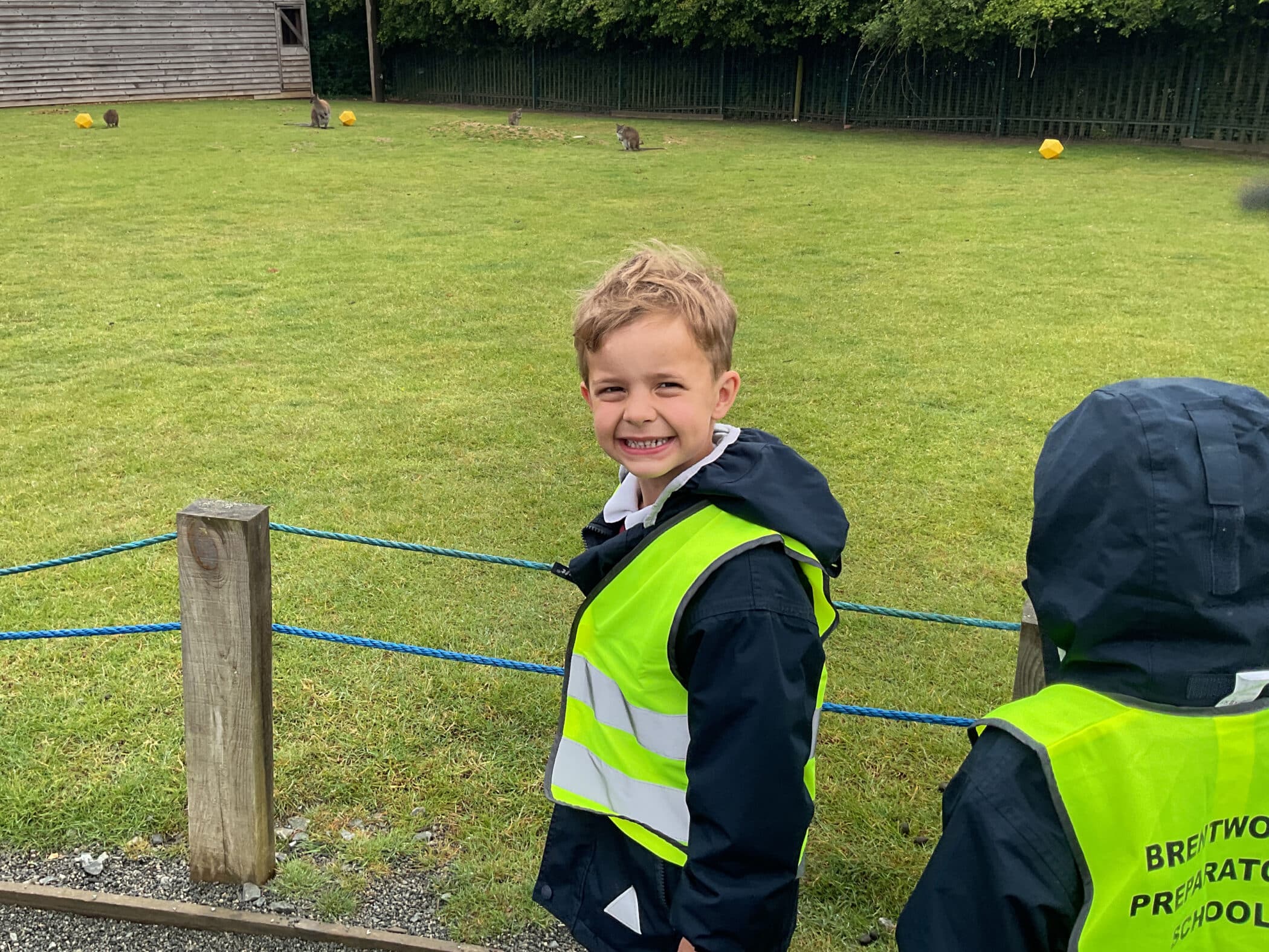 A young boy in a neon yellow safety vest smiles at the camera whilst standing next to a grassy field, accompanied by another child also wearing a vest. A wooden fence and small building are visible in the background.
