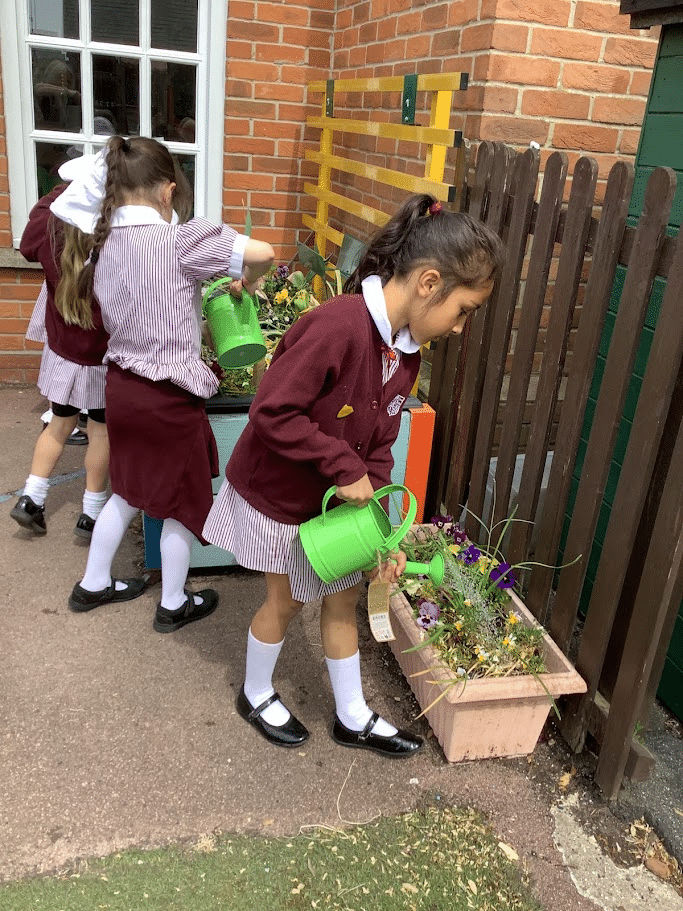 Three young girls in school uniforms water plants in rectangular pots by a fence outside, using green watering cans. The scene takes place near a brick building.
