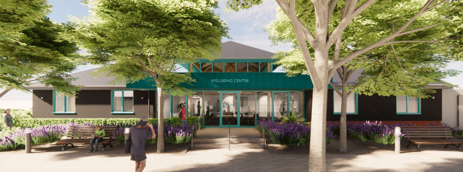 A modern building labelled Wellbeing Centre with large windows, surrounded by trees, benches, and purple flowers; a few people walk and sit in the shaded, landscaped outdoor area.