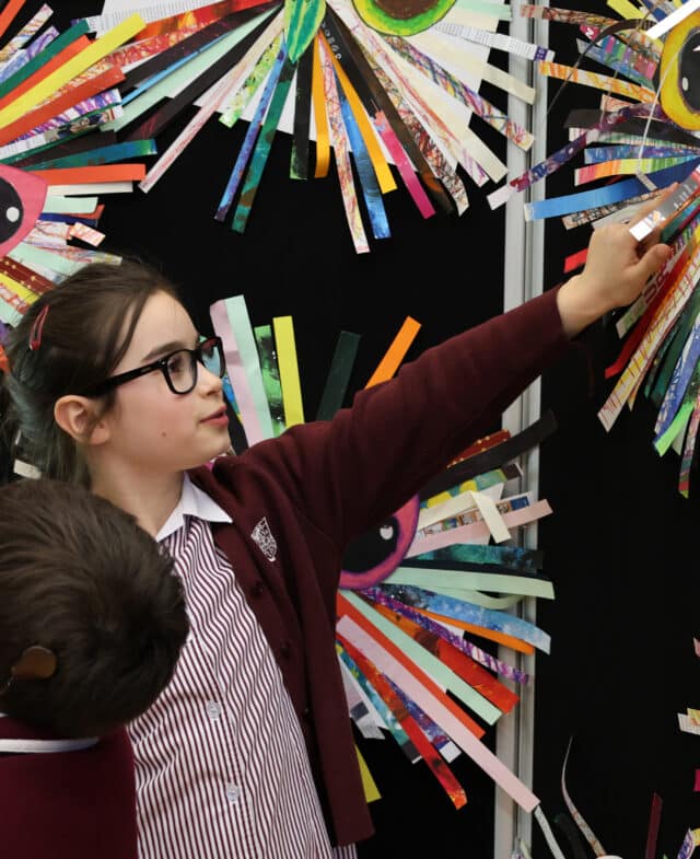A girl in glasses and a school uniform points at colourful, spiky, circular art on a wall—an engaging moment that highlights cultural capital—whilst another child stands nearby, partially visible in the foreground.