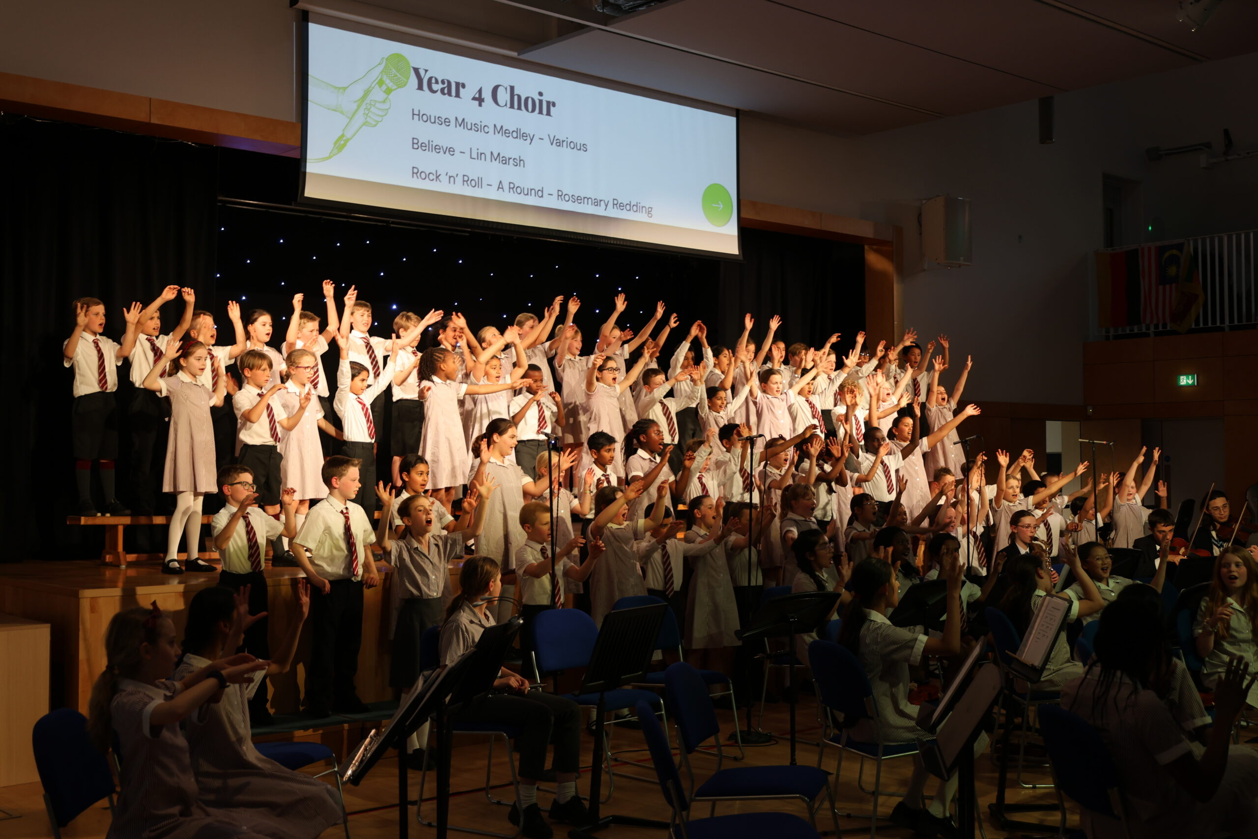 A large group of children in school uniforms stand on stage singing with raised arms, performing as the Year 4 Choir. A projector above displays their setlist to an audience in a school hall.