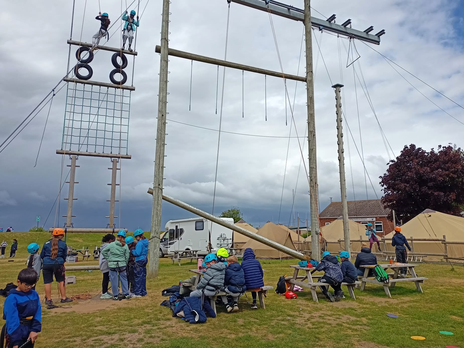 A group of children wearing helmets and harnesses gather outdoors near a high ropes course. Some kids climb the tall structure whilst others wait or sit at picnic tables. Tents and a caravan are in the background.