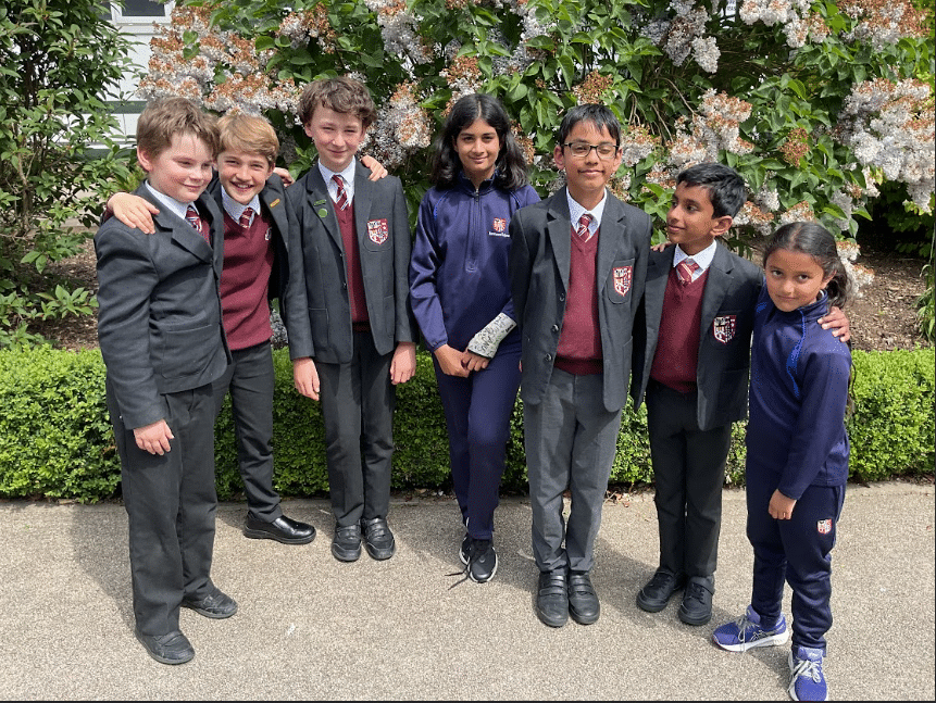 A group of seven children stand together outdoors in school uniforms, smiling in front of flowering bushes and greenery. Six wear blazers and jumpers; one girl wears a navy tracksuit.