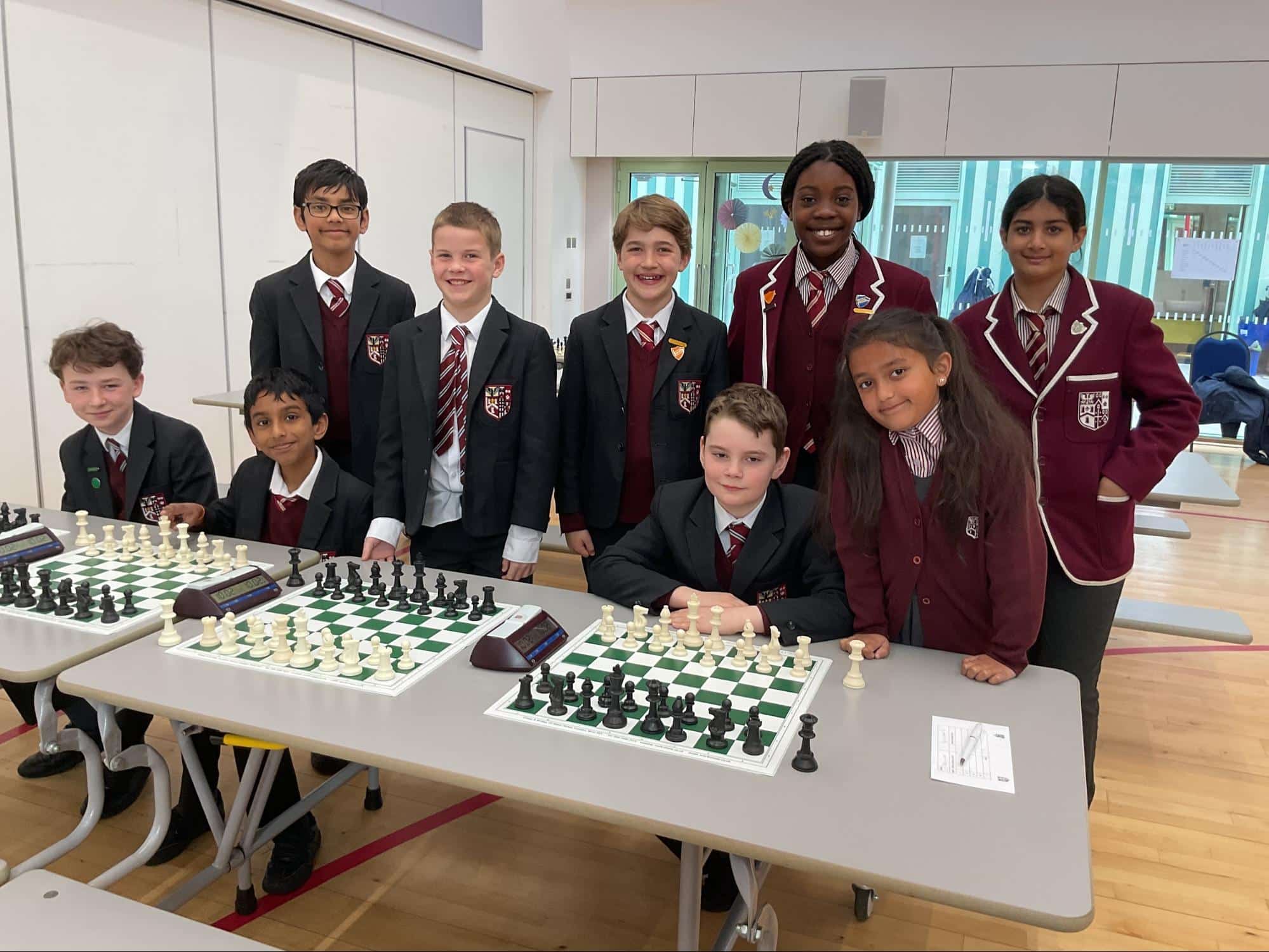 A group of schoolchildren in uniforms pose and smile around tables with chessboards in a bright indoor setting, ready for a chess competition or club activity.
