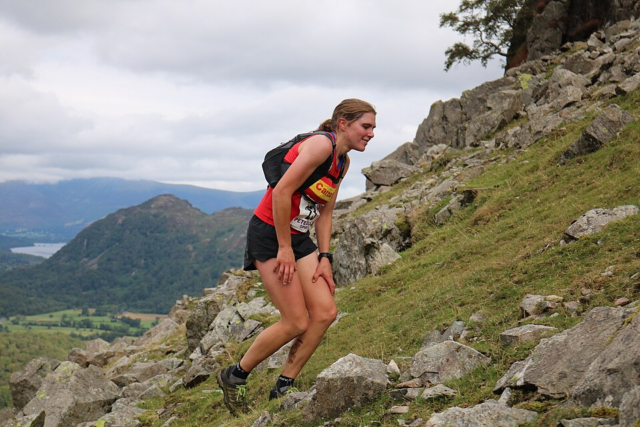 A runner wearing a red shirt and black shorts climbs a steep, rocky hillside with a rucksack, appearing tired. Mountains and a lake are visible in the background under a cloudy sky.