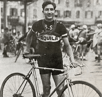 A smiling male cyclist in vintage racing attire stands outdoors holding a bicycle. Other cyclists and blurred buildings appear in the background. His jersey reads S.S. AQUILA.