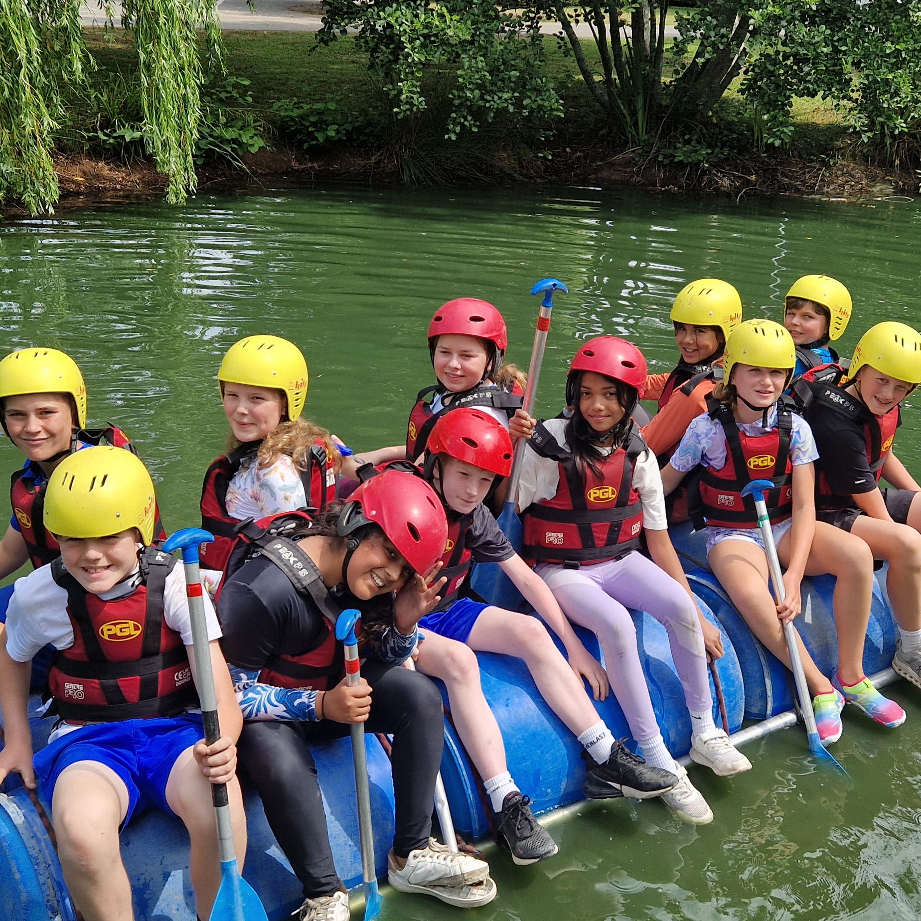 A group of children wearing helmets and life jackets sit on a homemade raft made of blue barrels, smiling and holding paddles on a green lake, surrounded by trees.