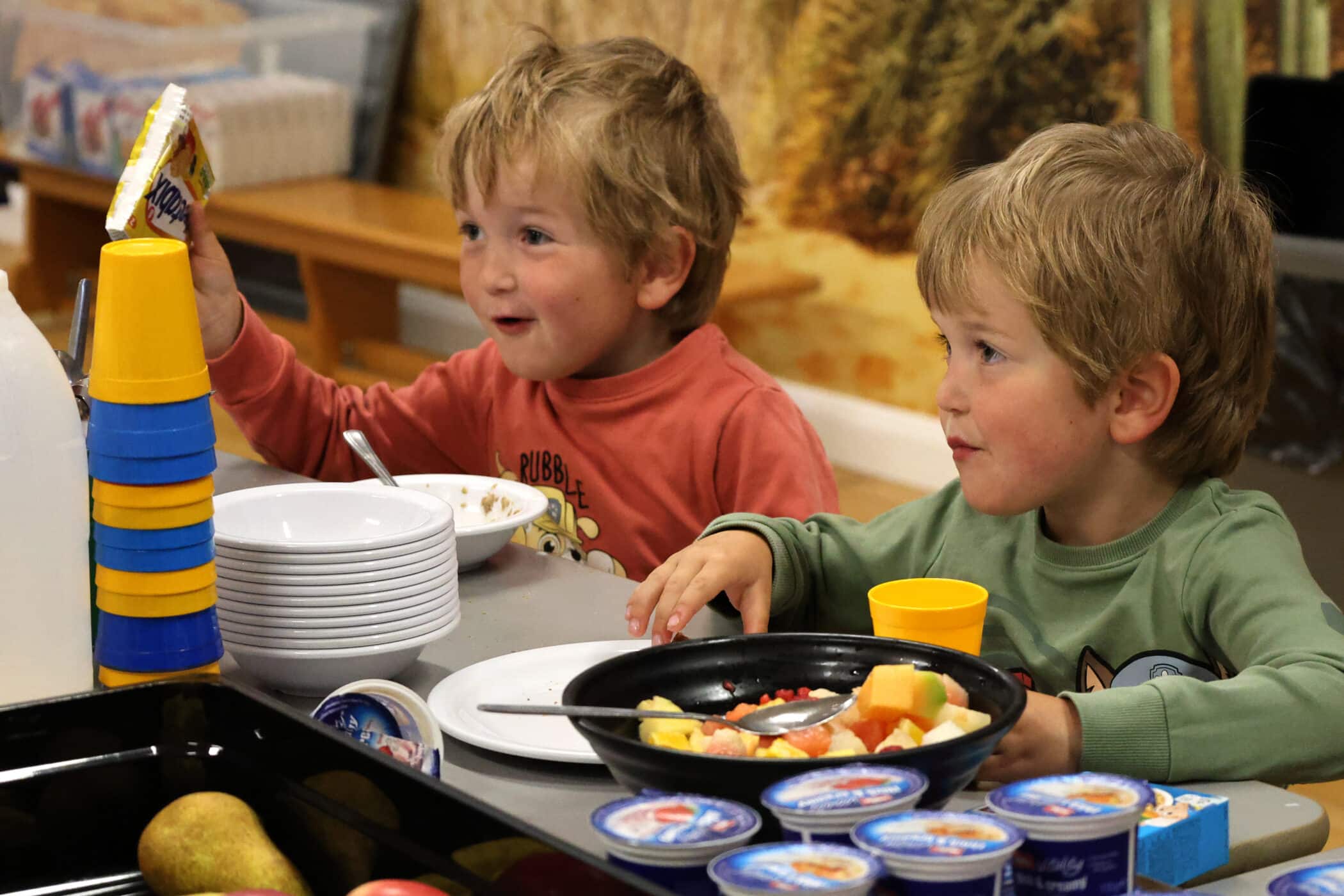 Two young children sit at a table eating breakfast. They have bowls, cups, yoghurt, and a plate of fruit in front of them. One child is smiling and holding up a snack, while the other looks ahead with a slight smile.