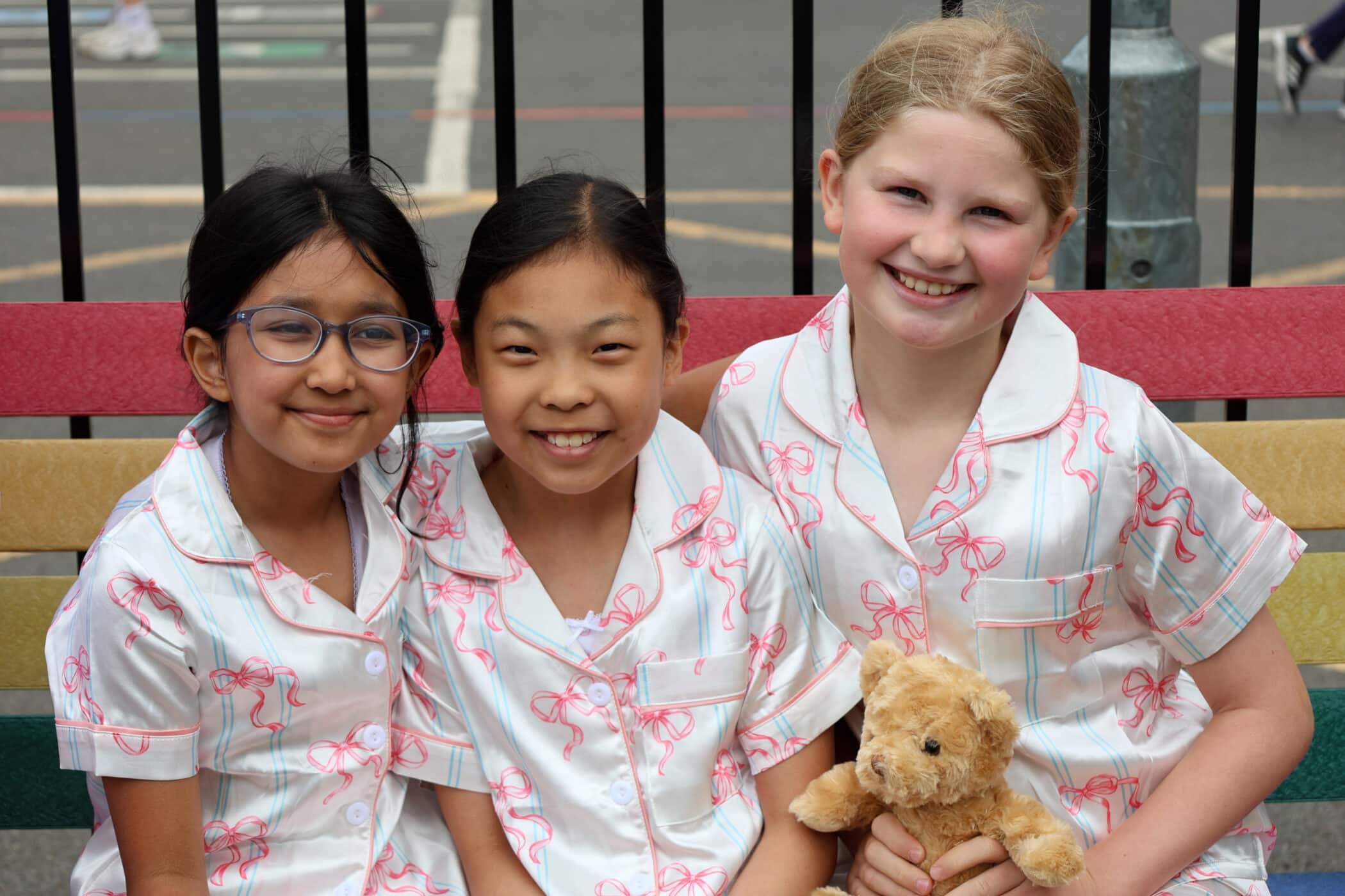Three smiling girls in matching white pyjamas with pink prints sit on a colourful bench. One holds a small plush teddy bear. They are outdoors, with a metal fence and playground in the background.