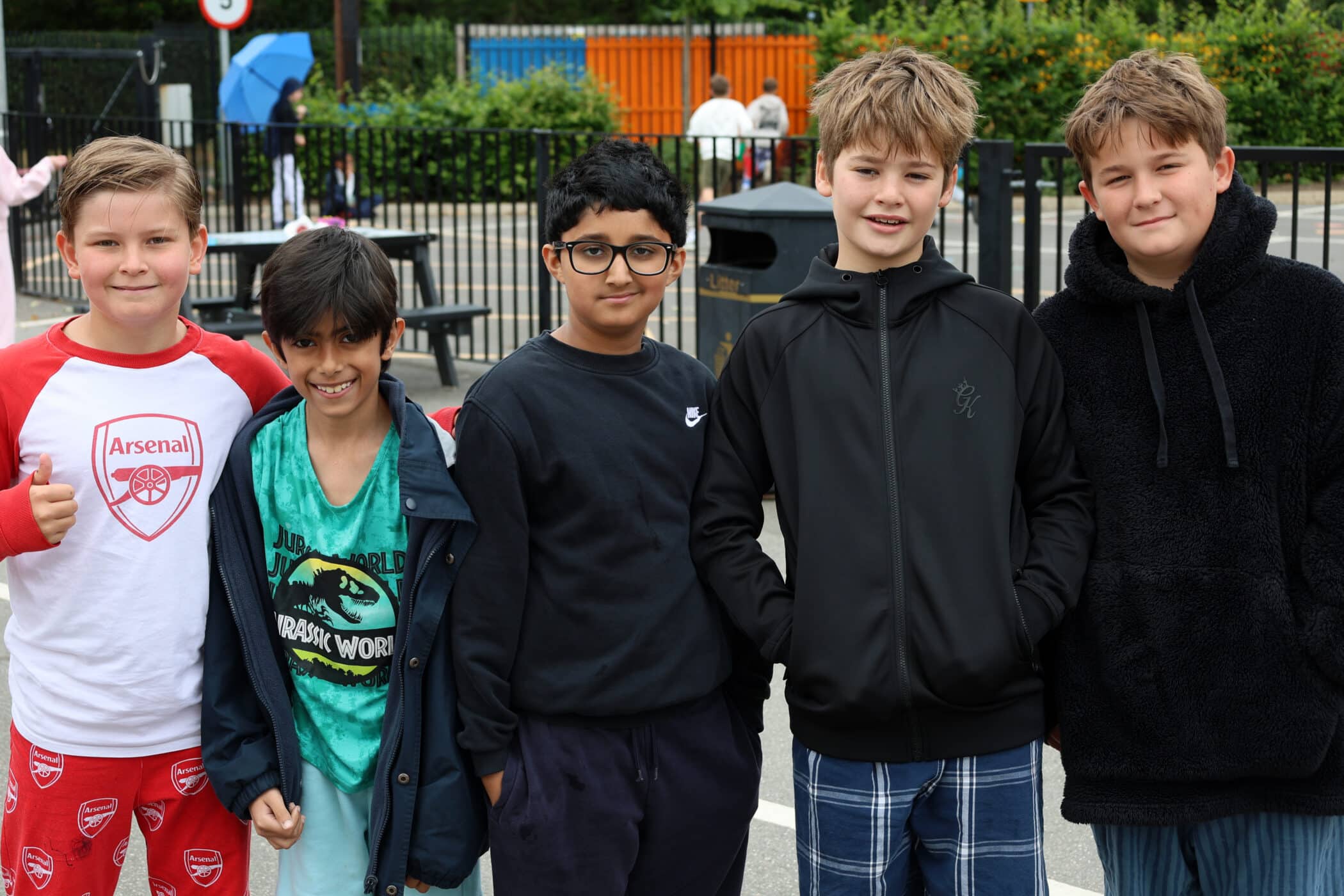 Five boys stand side by side outdoors, smiling and posing for the camera. They are dressed casually in shorts, T-shirts, and jackets, with a playground and trees visible in the background.