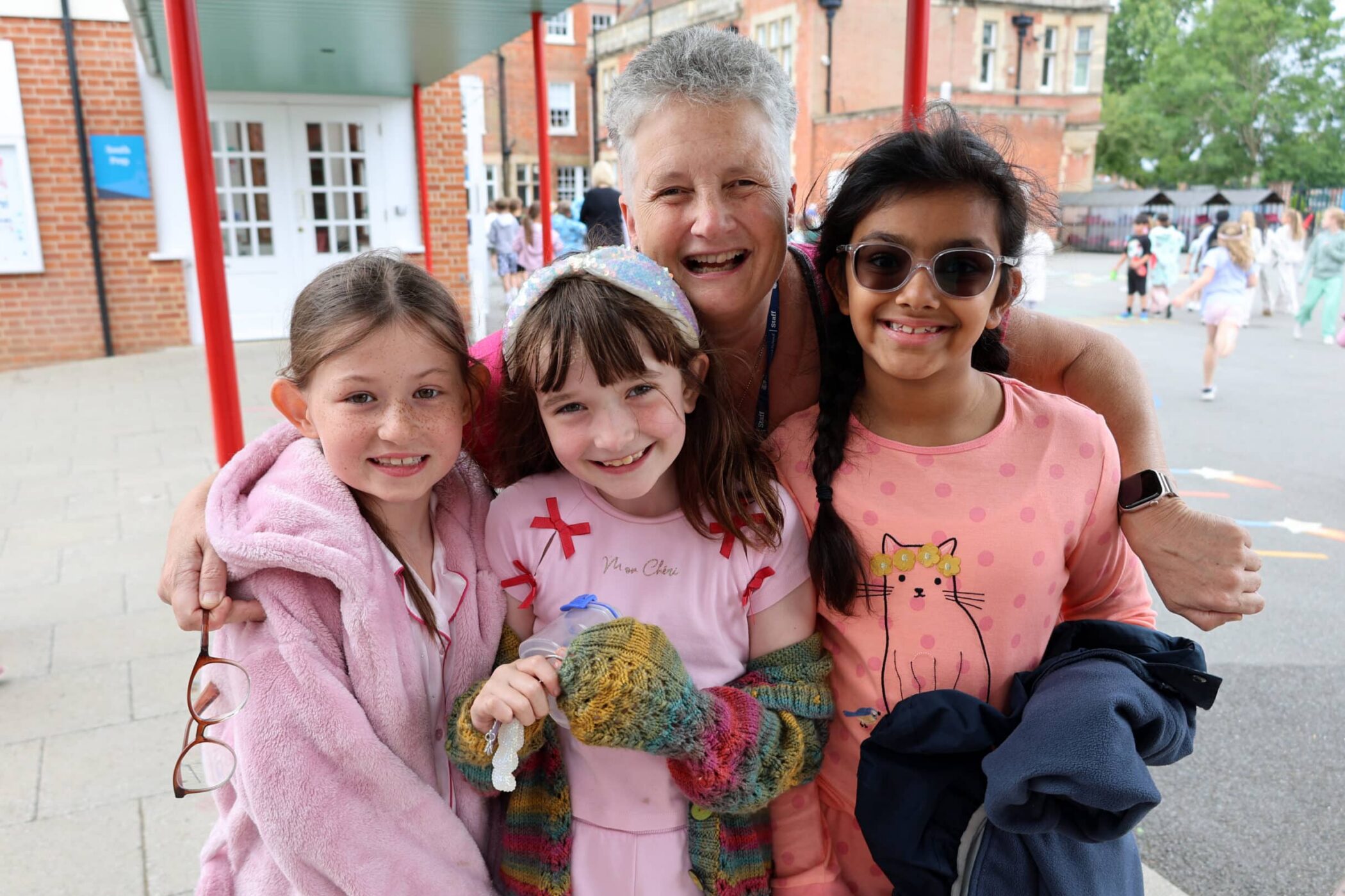 An older woman hugs three smiling young girls outdoors at a school. The girls wear light, colourful clothes and glasses, and the group looks happy. Other children and school buildings appear in the background.