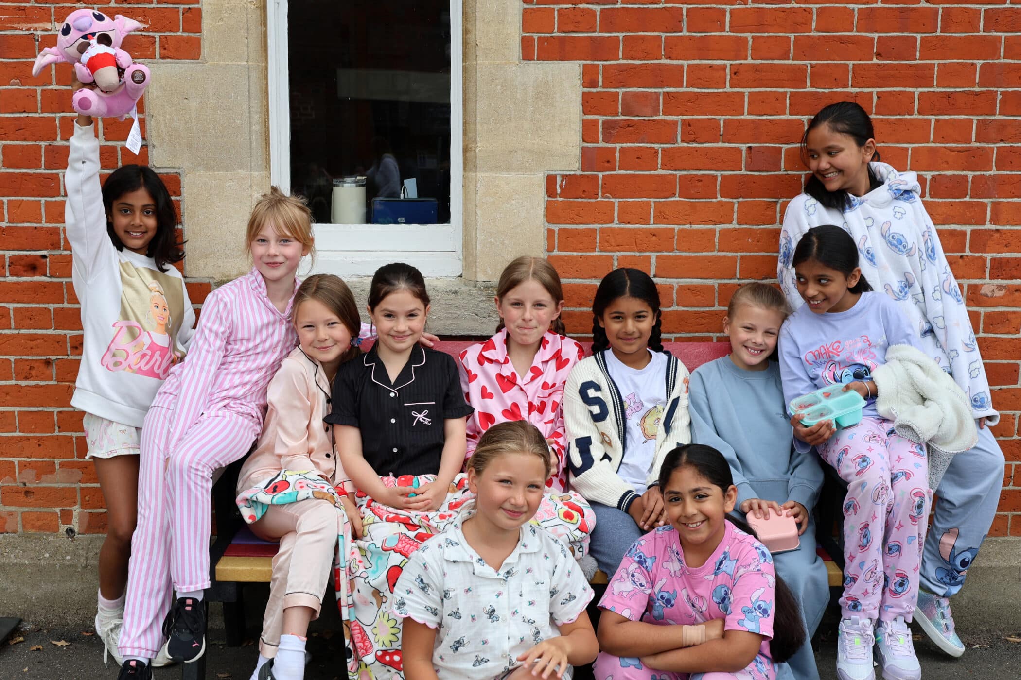 A group of eleven smiling girls wearing colourful pyjamas pose together outside in front of a brick wall, with one girl on the left holding up a pink soft toy.