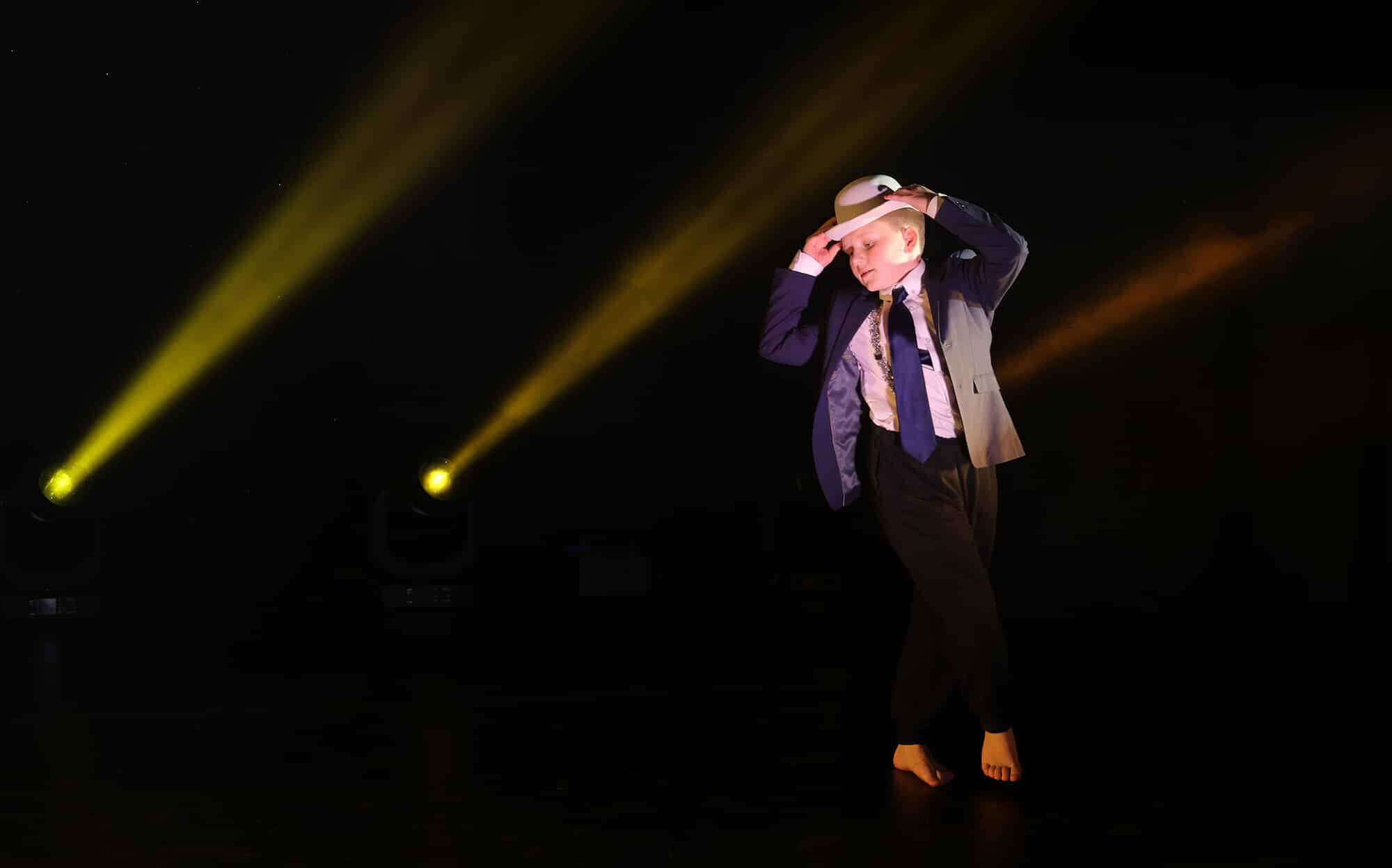 A barefoot dancer in a suit, tie, and hat performs on a dark stage with dramatic yellow spotlights shining in the background, embodying the dreams and ambitions scholarships help make possible.