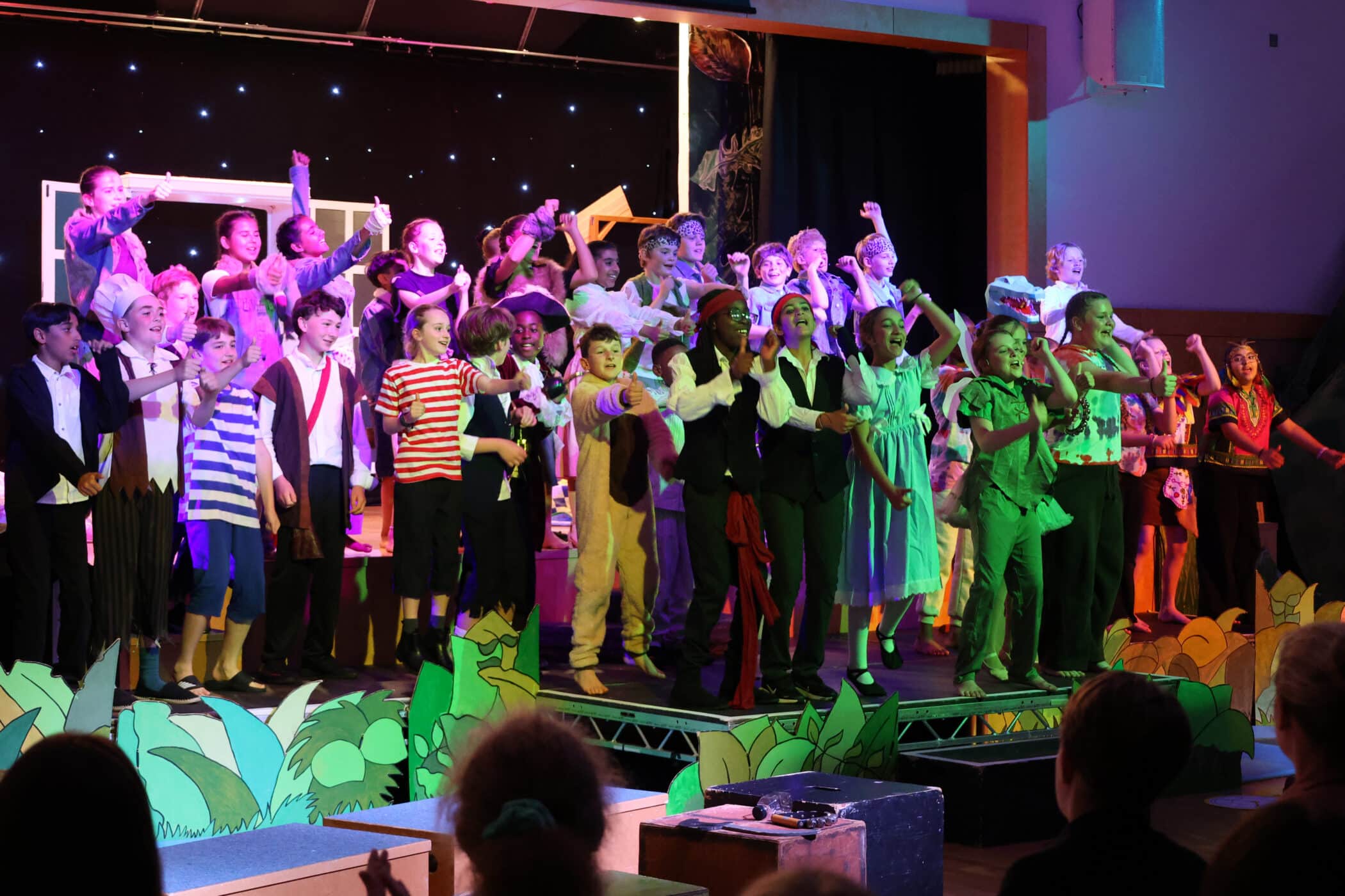 A large group of children in colourful costumes perform on stage during a play, gesturing enthusiastically. The set includes jungle plants, a starry night backdrop, and an audience watches from below.