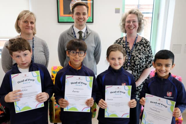 Four smiling children hold Head of Prep Award certificates, standing in front of three adults. All are indoors, posing for a group photo, with bright decor and a window in the background.
