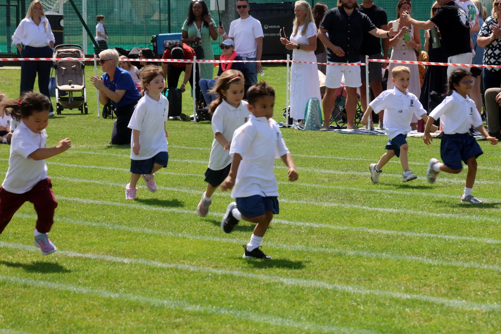 Young children in sports kit race on a grass track while adults watch from behind a barrier. The scene is outdoors, sunny, and lively, with the audience standing or sitting in the background.