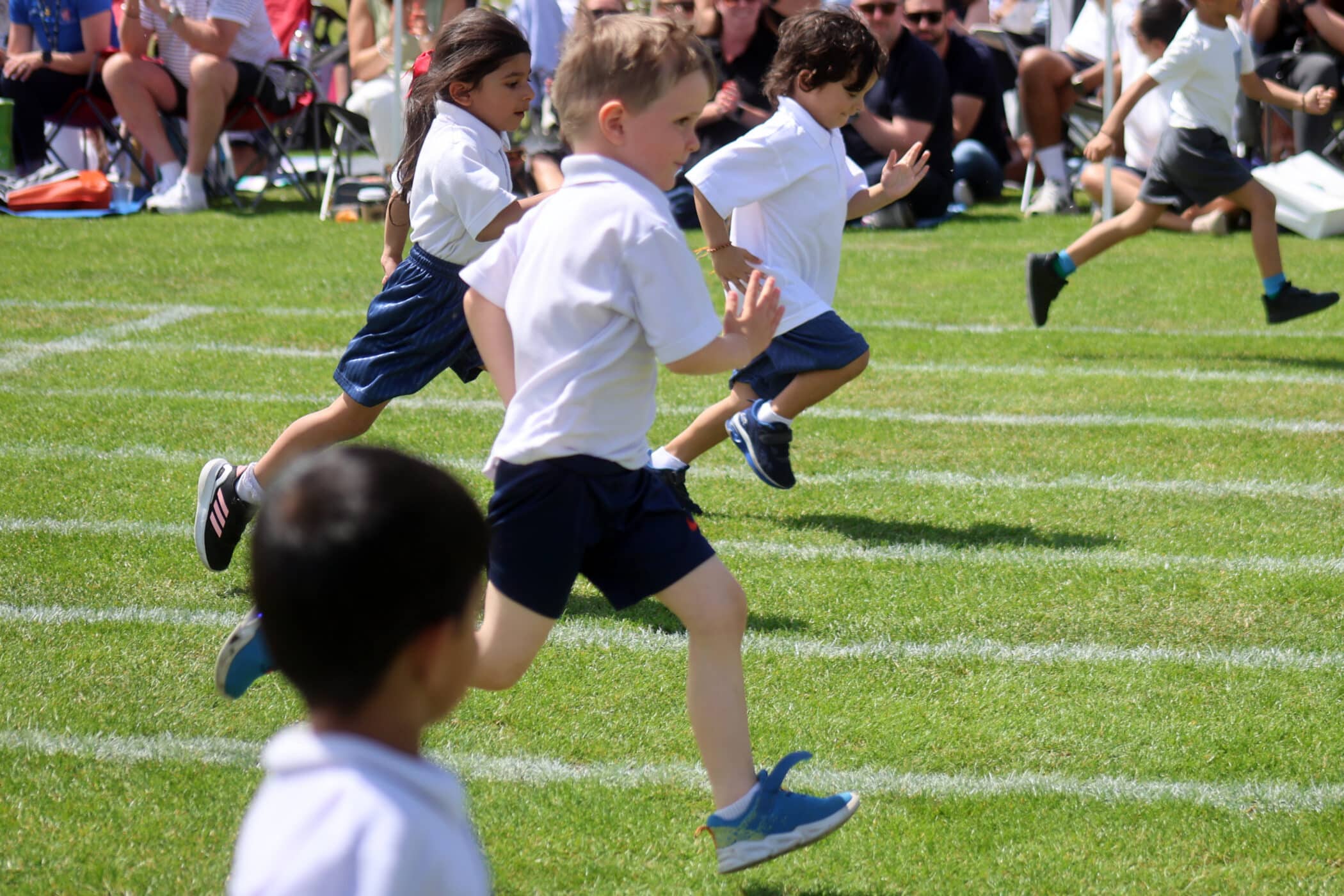 Children wearing sports kits are running a race on a grassy track, with spectators seated in the background watching the event. The scene is lively, with children in motion under a sunny sky.