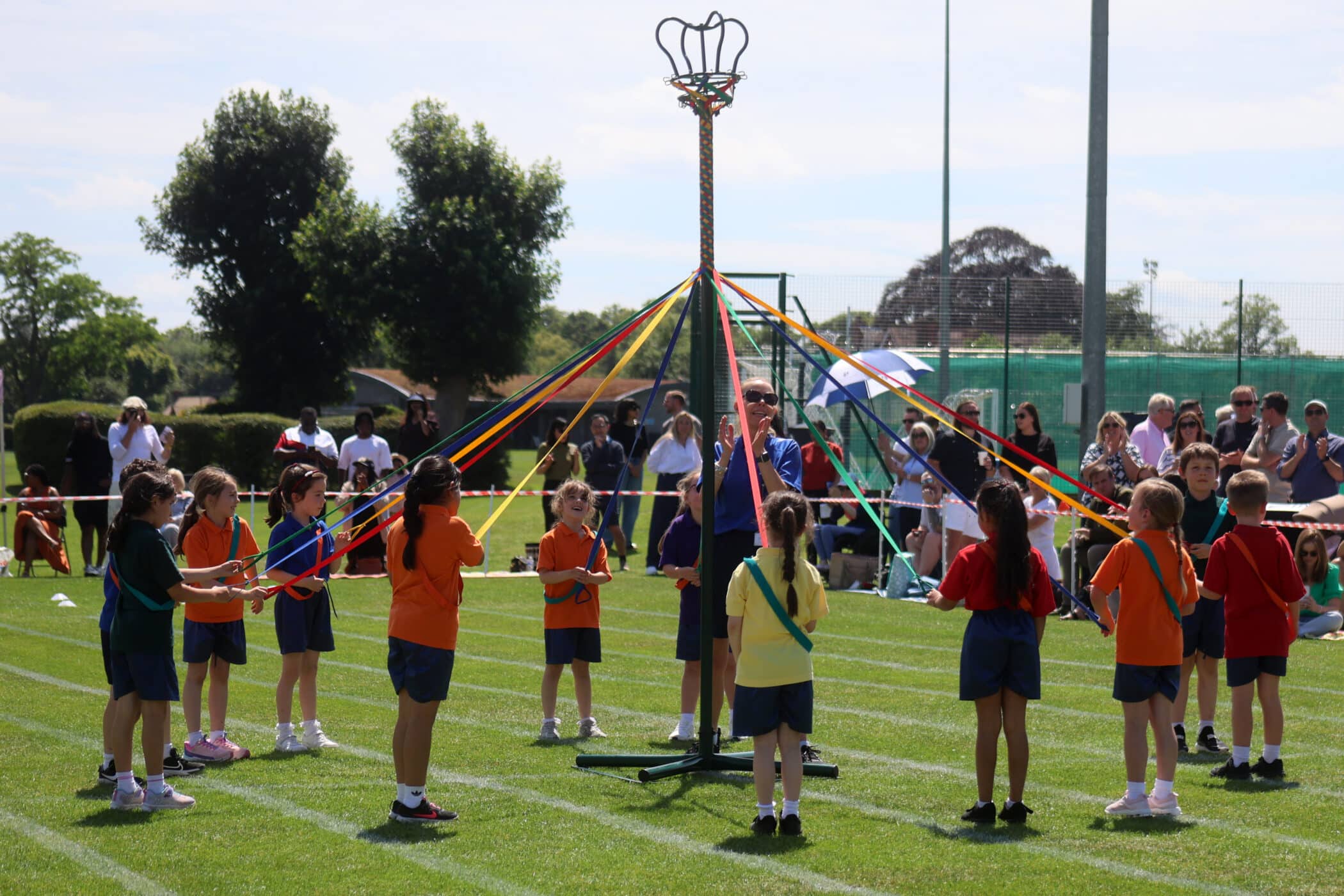 Children in colourful outfits hold ribbons and dance around a maypole on a grassy field during a school event, with spectators watching in the background under sunny skies.