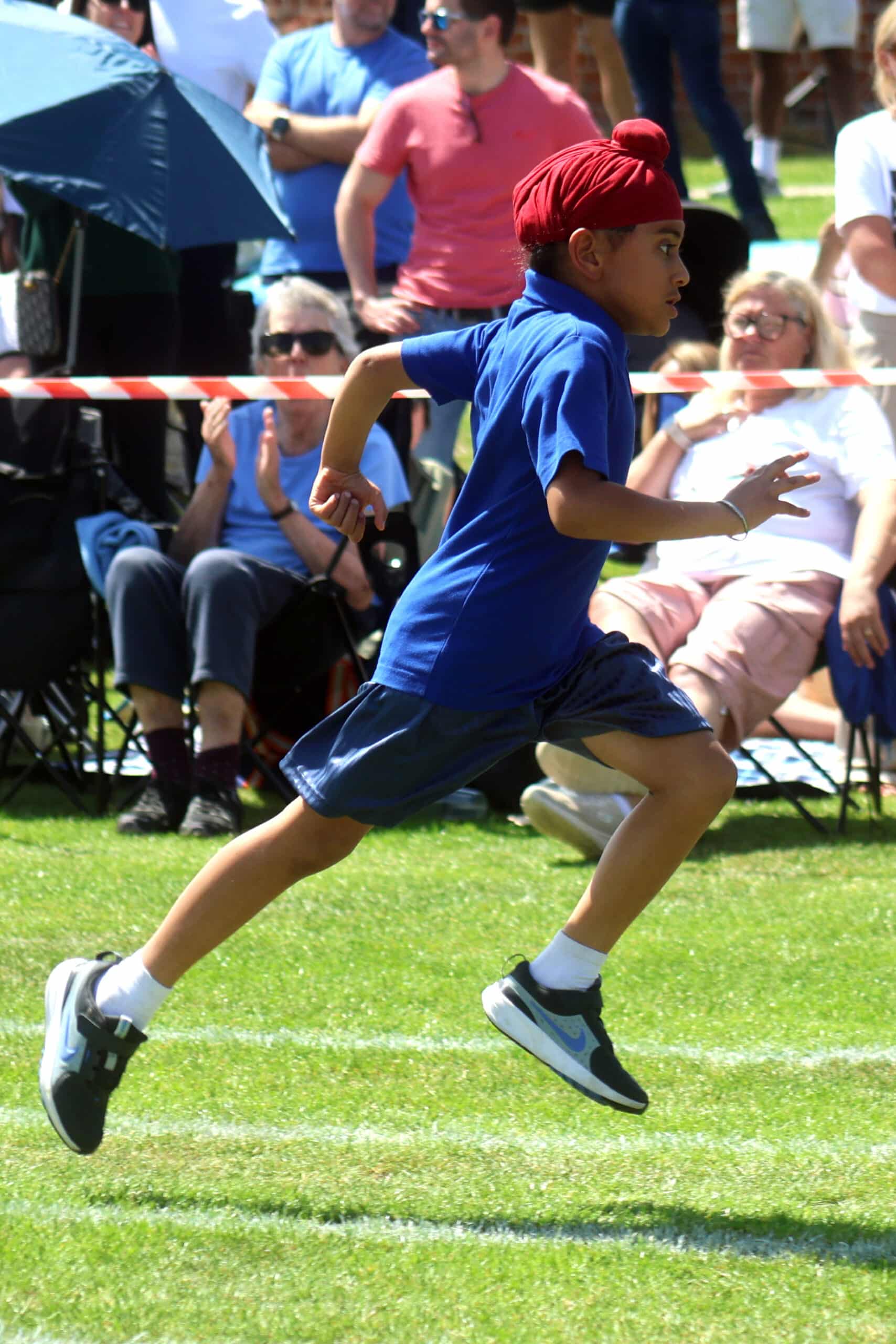 A young boy wearing a red turban, blue shirt, and shorts runs in a race on a grassy field, with spectators seated and standing in the background on a sunny day.