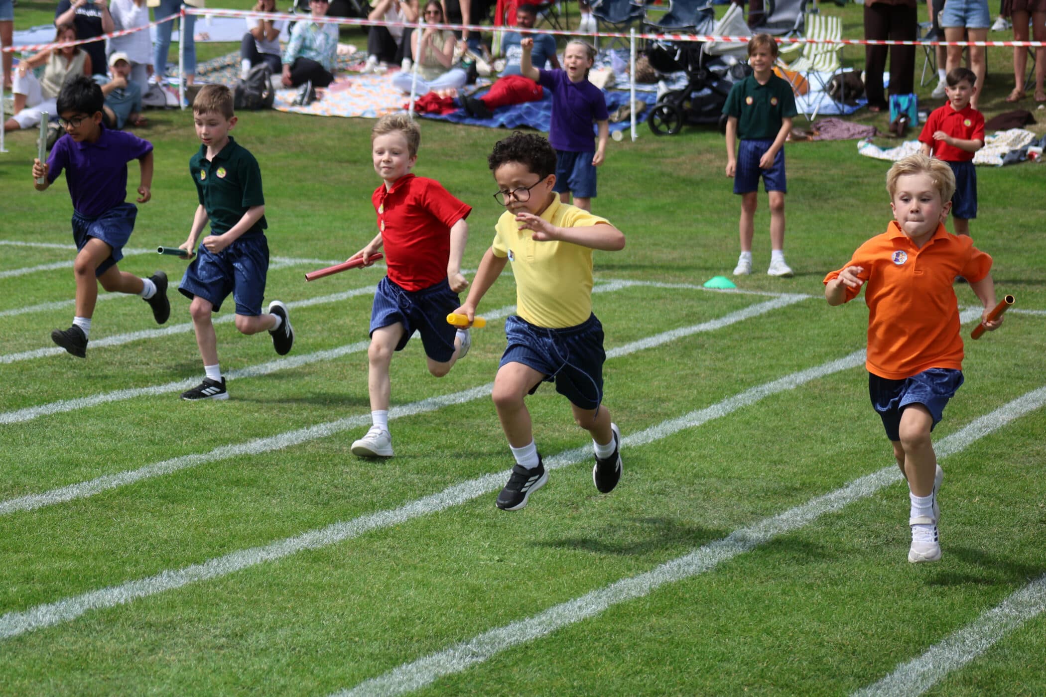 A group of young children race on a grassy track, each holding a relay baton. Spectators sit and watch in the background on a sunny day.