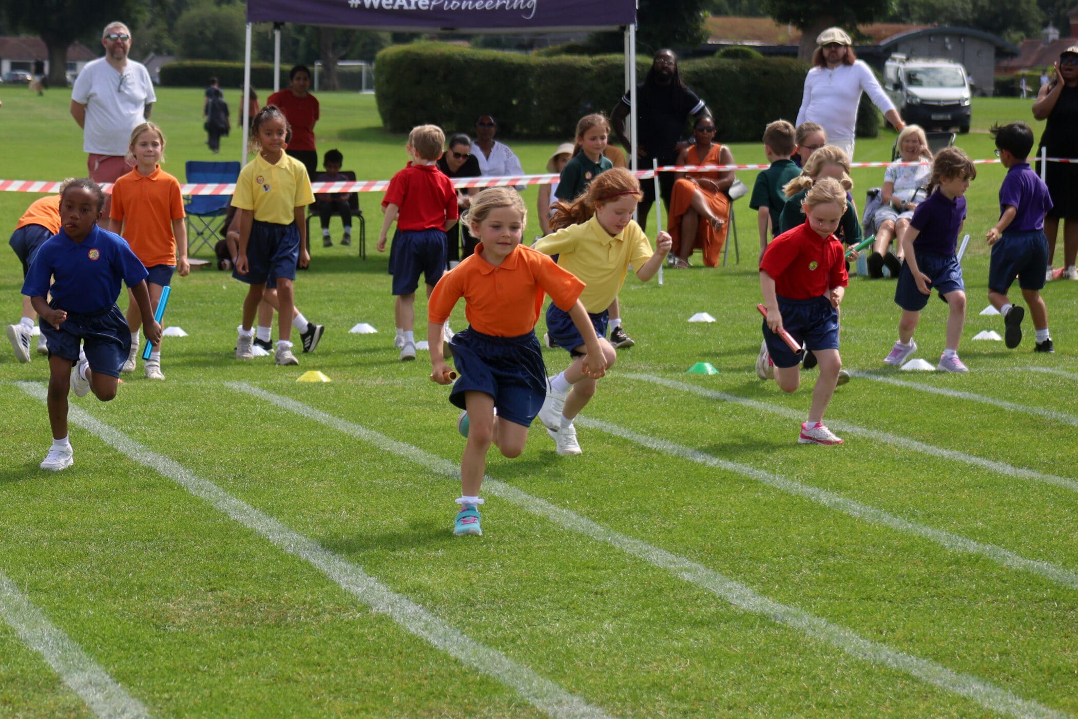 A group of young children in colourful shirts sprinting on a grassy field during a race, with adults and other children watching in the background. Lines and cones mark the race lanes.