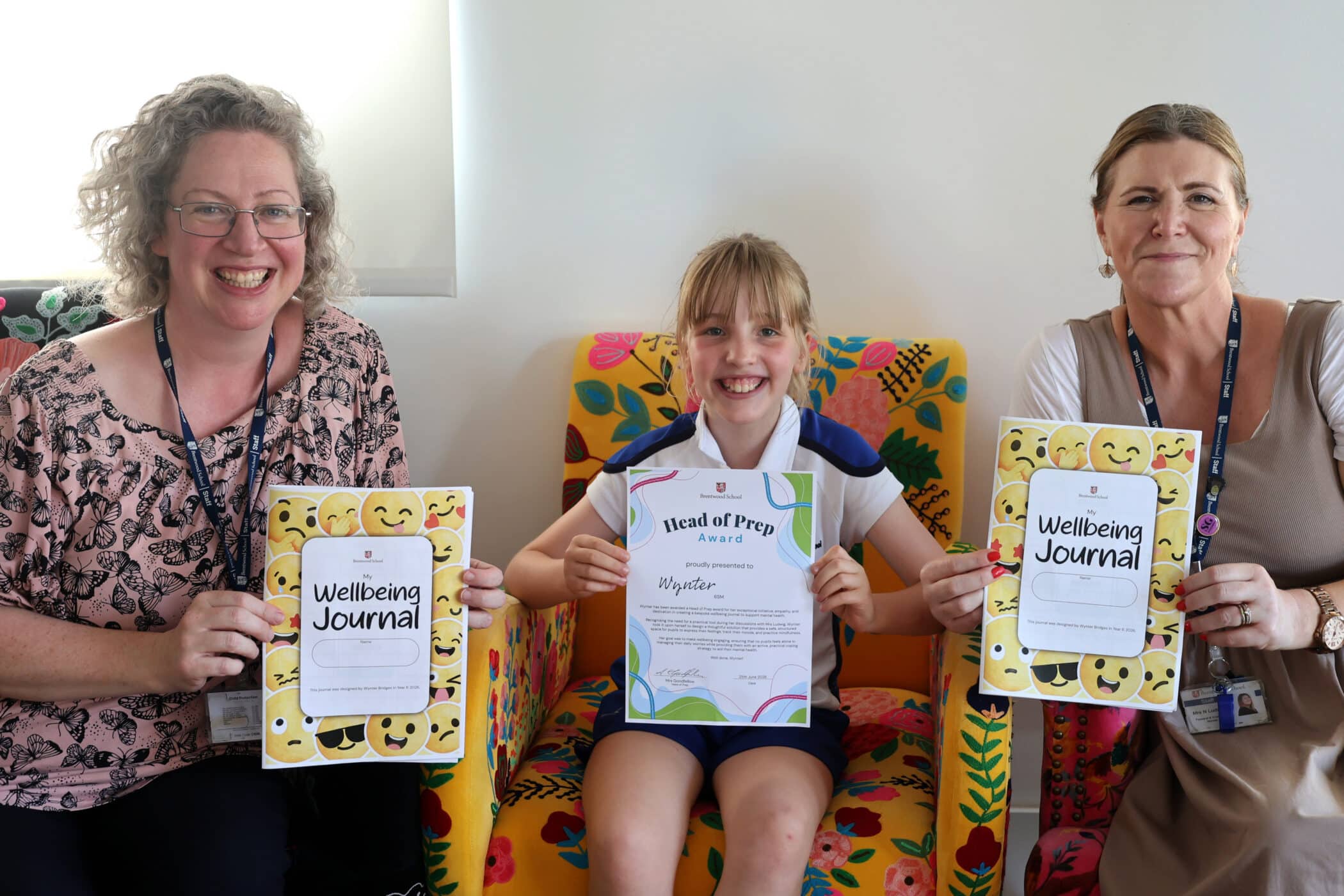 A smiling girl sits between two women on colourful chairs, holding an award certificate. The women each hold “Wellbeing Journal” booklets with emoji covers. All appear happy and proud.