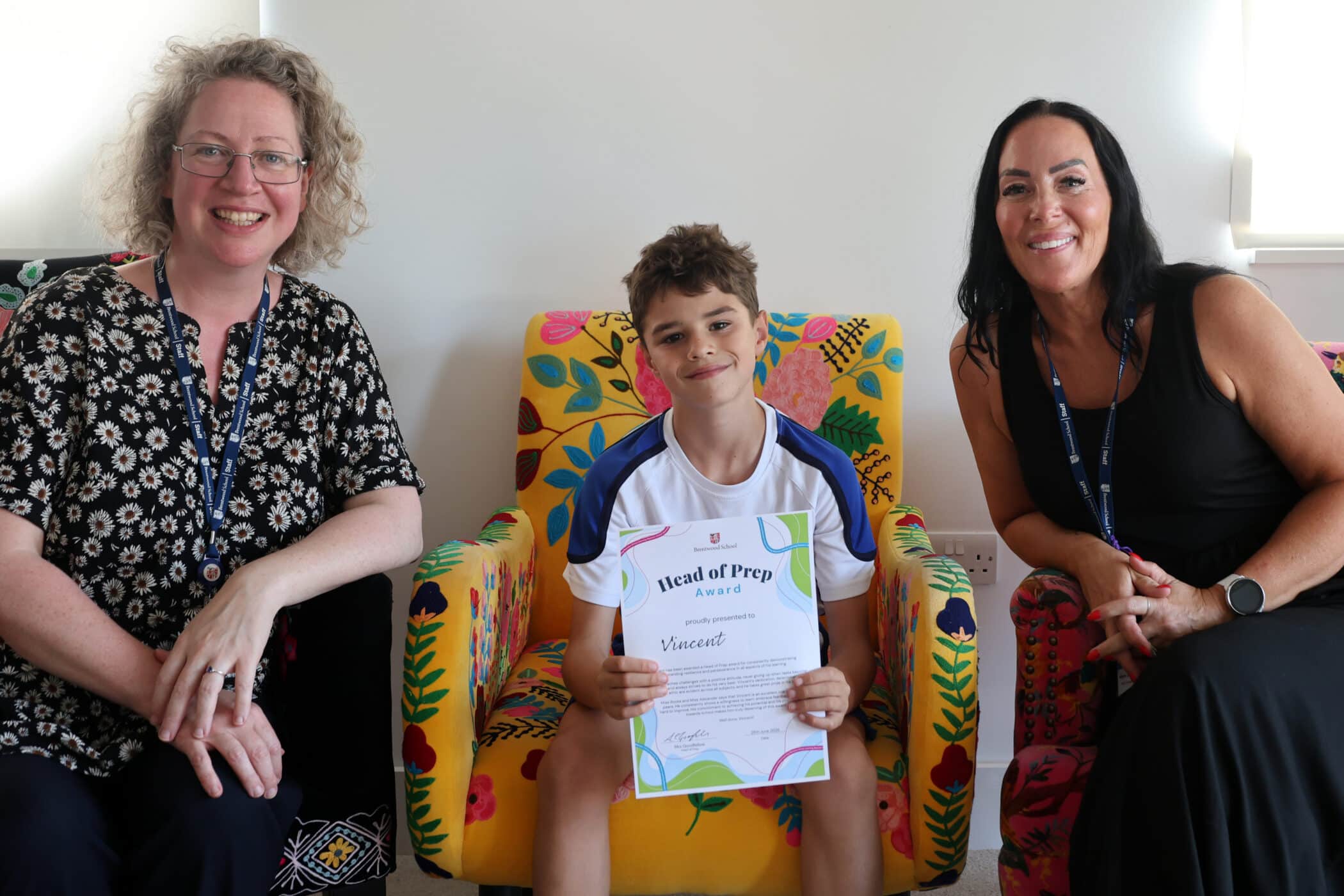 A smiling child sits between two women on colourful chairs, proudly holding a Head of Prep Award certificate. All three look happy, and the background includes vibrant, floral-patterned upholstery.